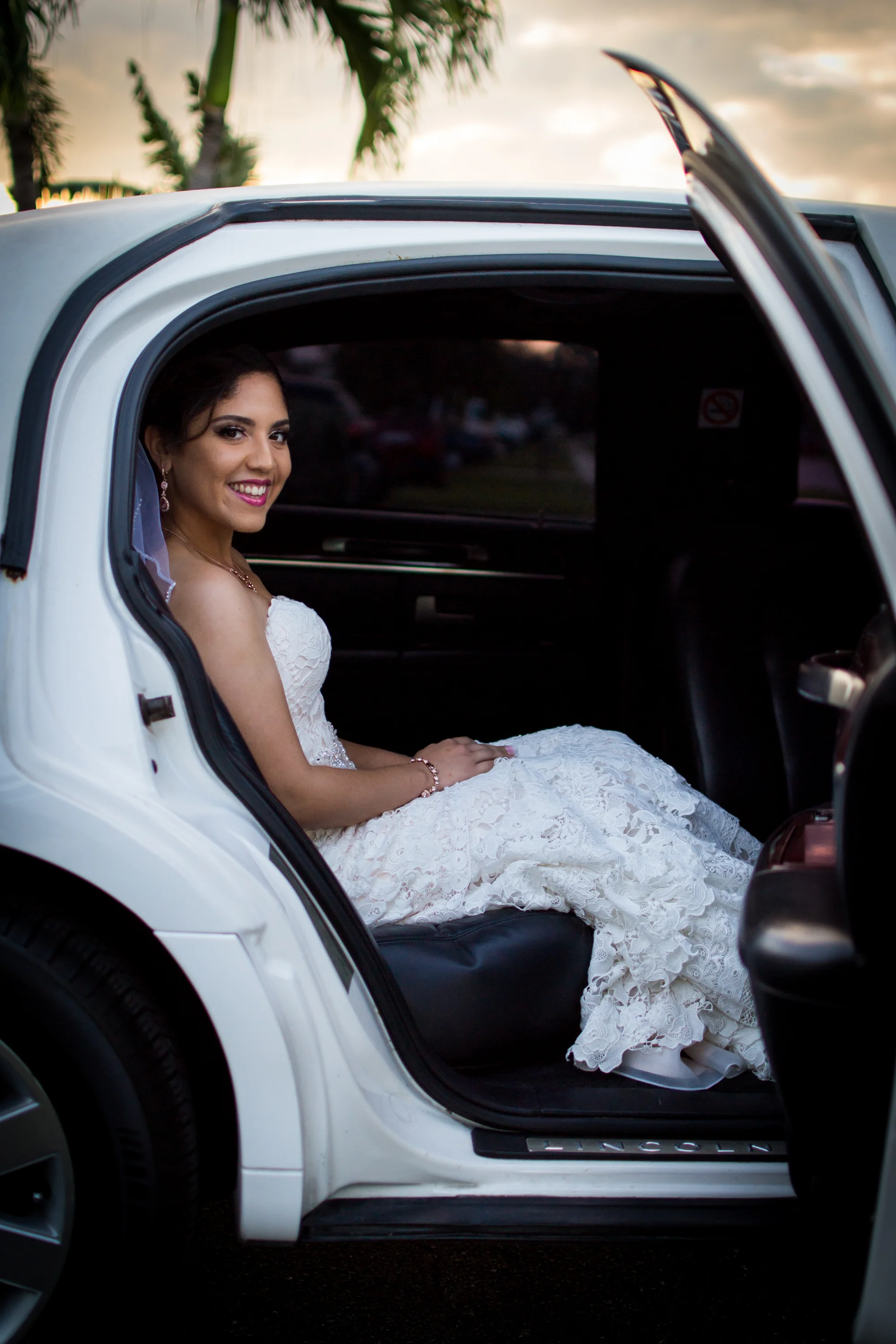 Bride in a white dress sitting in a white limousine with an open door, smiling, with a sunset background.
