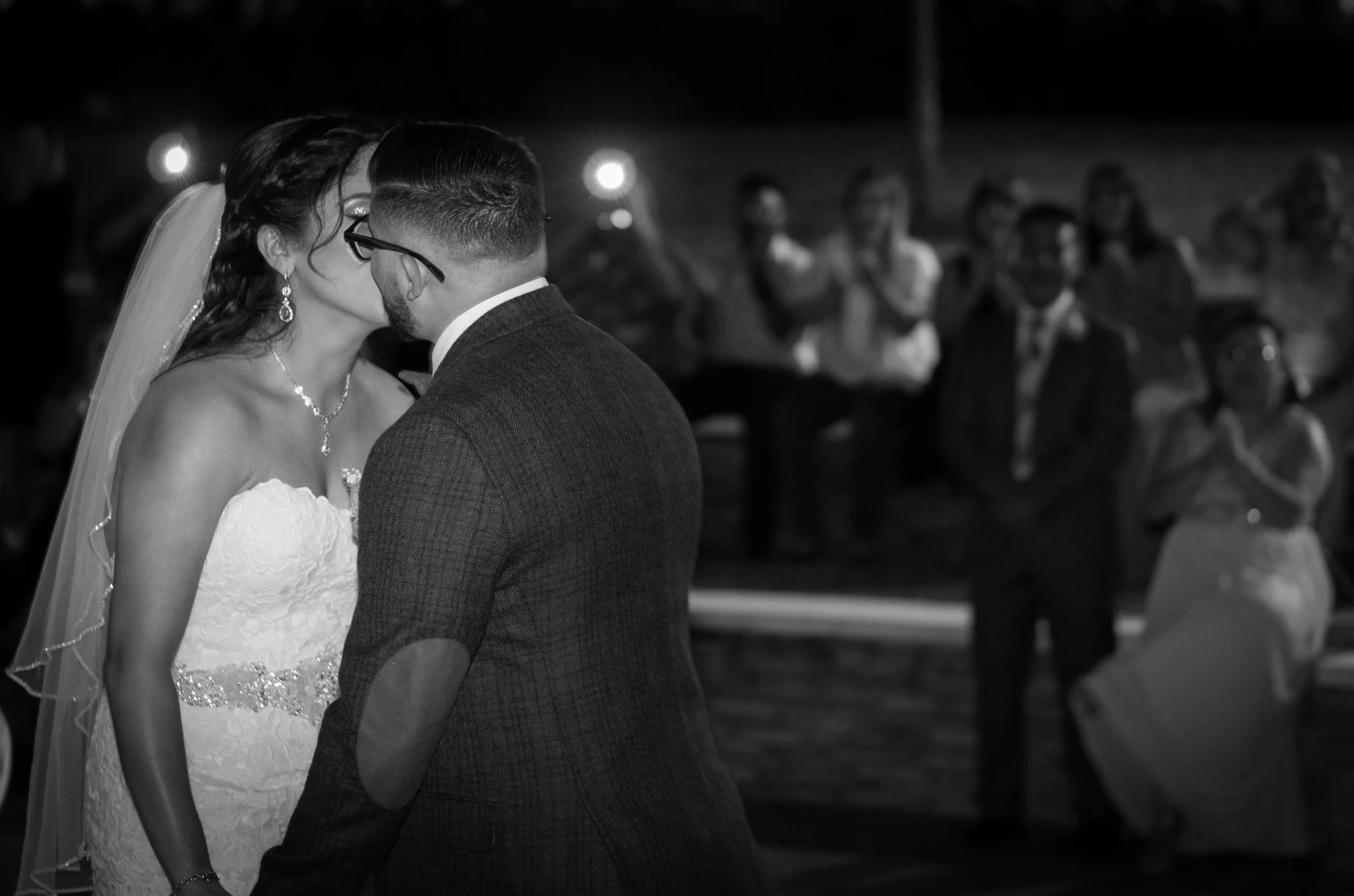 Black and white wedding photo of a bride and groom kissing, with a seated audience blurred in the background.