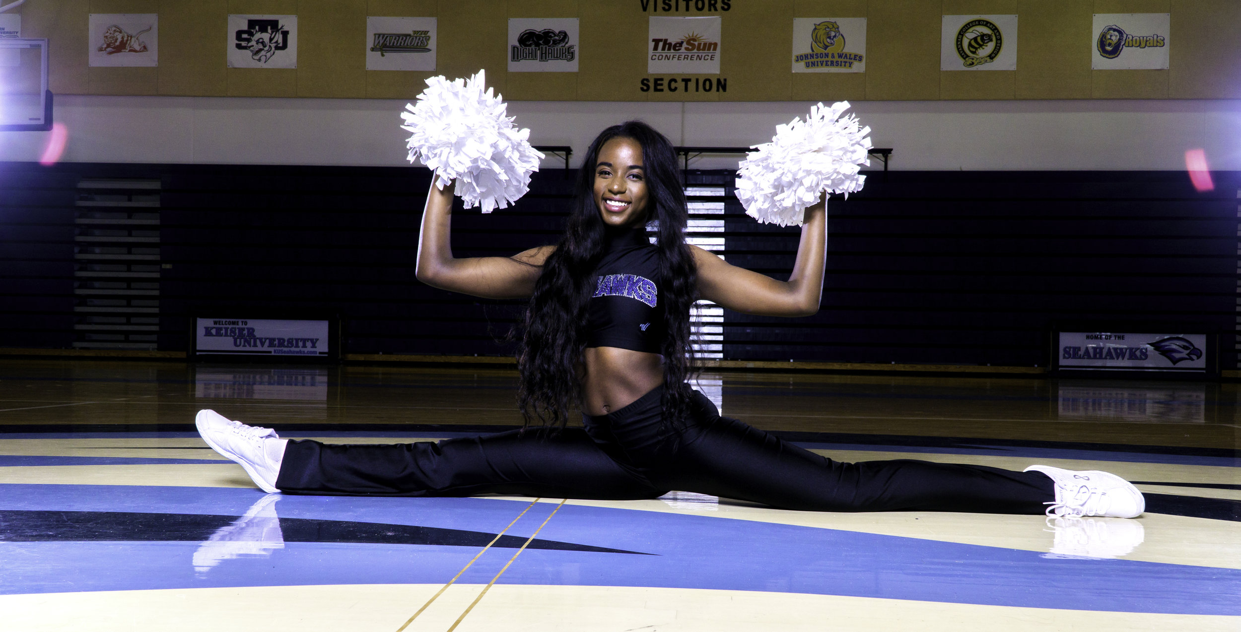 Cheerleader performing a split with pom-poms in a sports arena, wearing a black uniform and white sneakers.