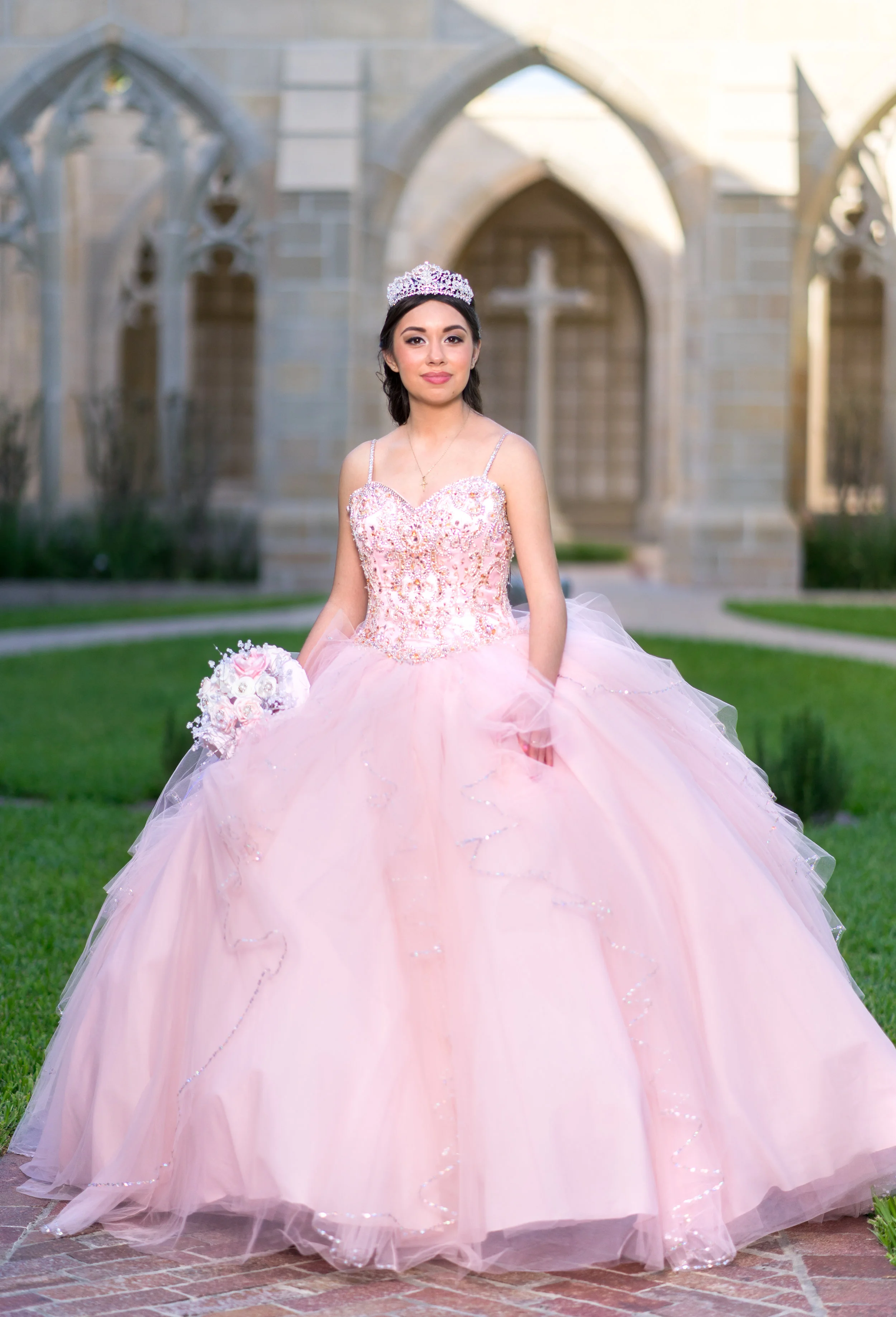 Young woman in pink quinceañera dress holding a bouquet, standing outdoors with an arched building background.