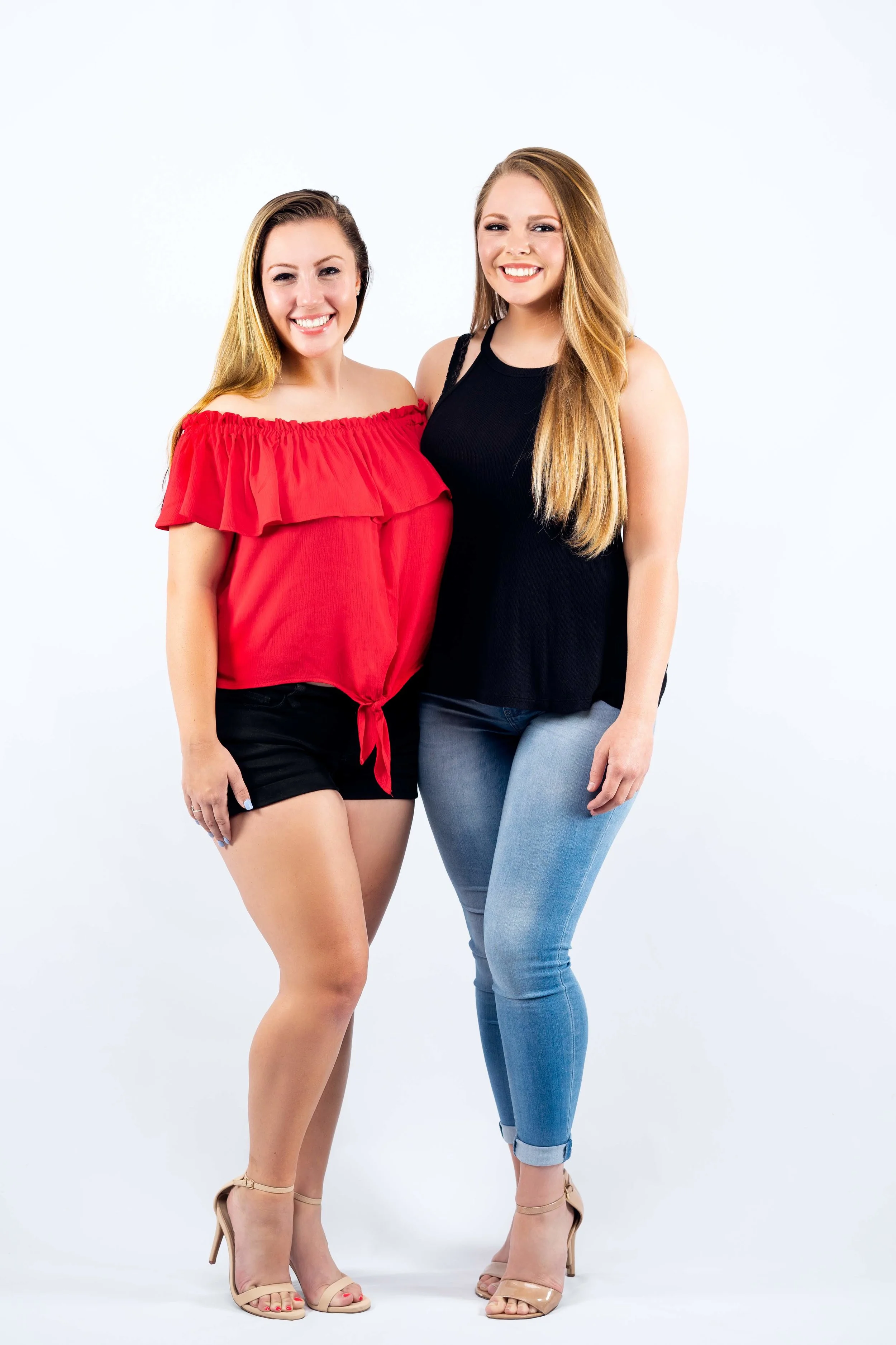Two smiling women, one in a red off-shoulder top and black shorts, and the other in a black sleeveless top and blue jeans, standing side by side.