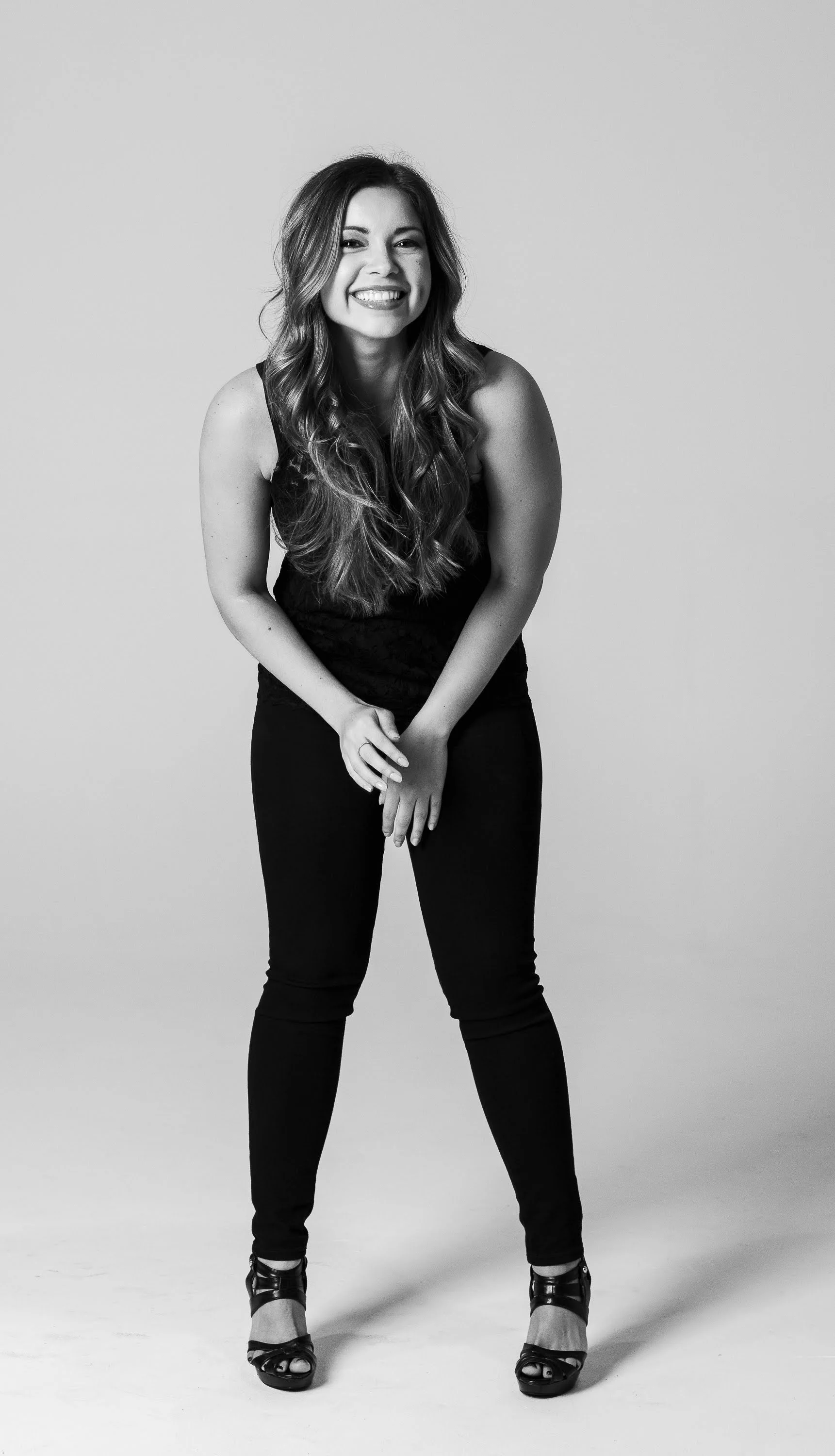 A woman smiling and leaning forward, wearing a black sleeveless top and black pants, posed against a plain background.