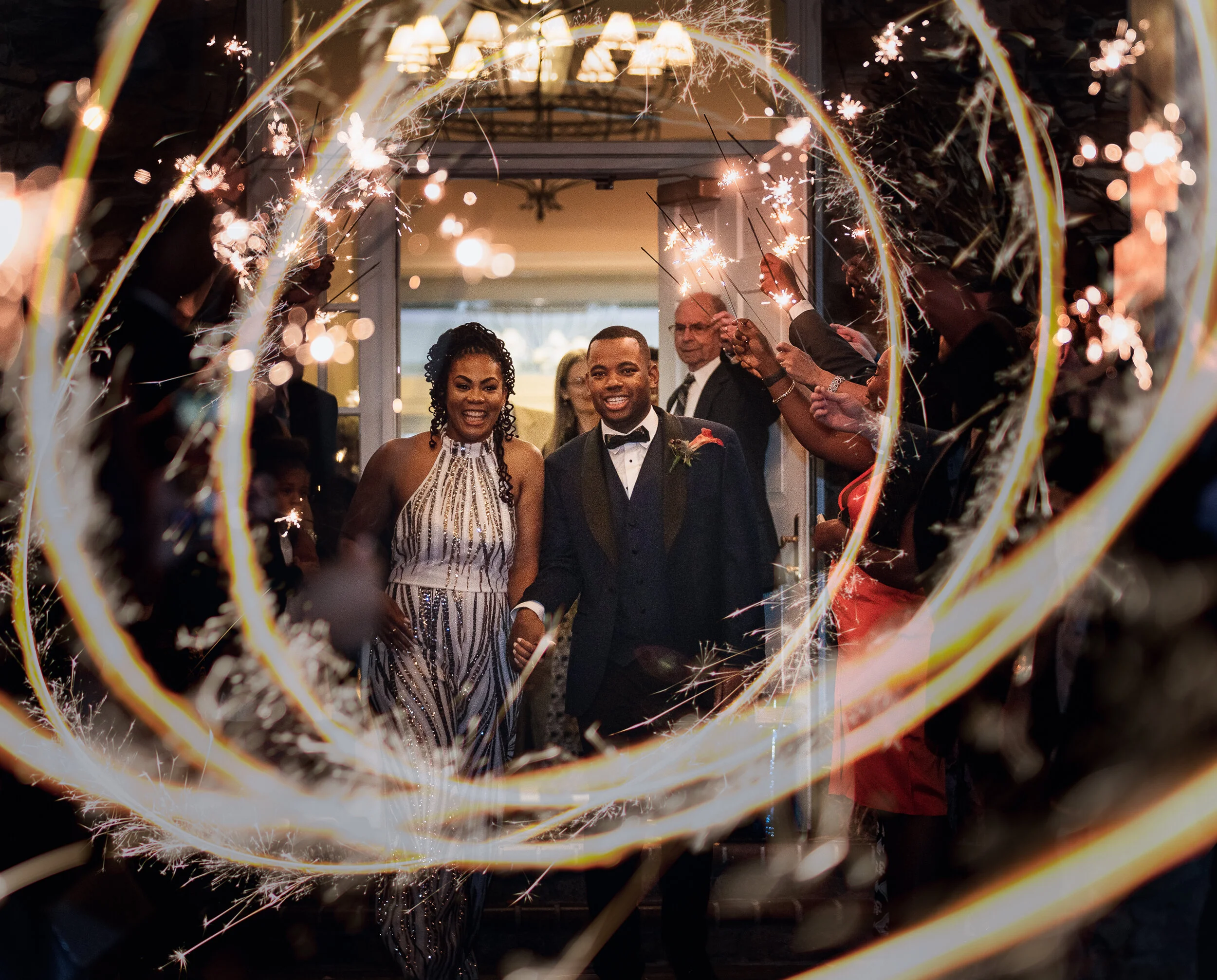 A couple dressed in formal attire walking through a sparkler exit at a wedding reception, surrounded by guests holding sparklers, creating a festive atmosphere.