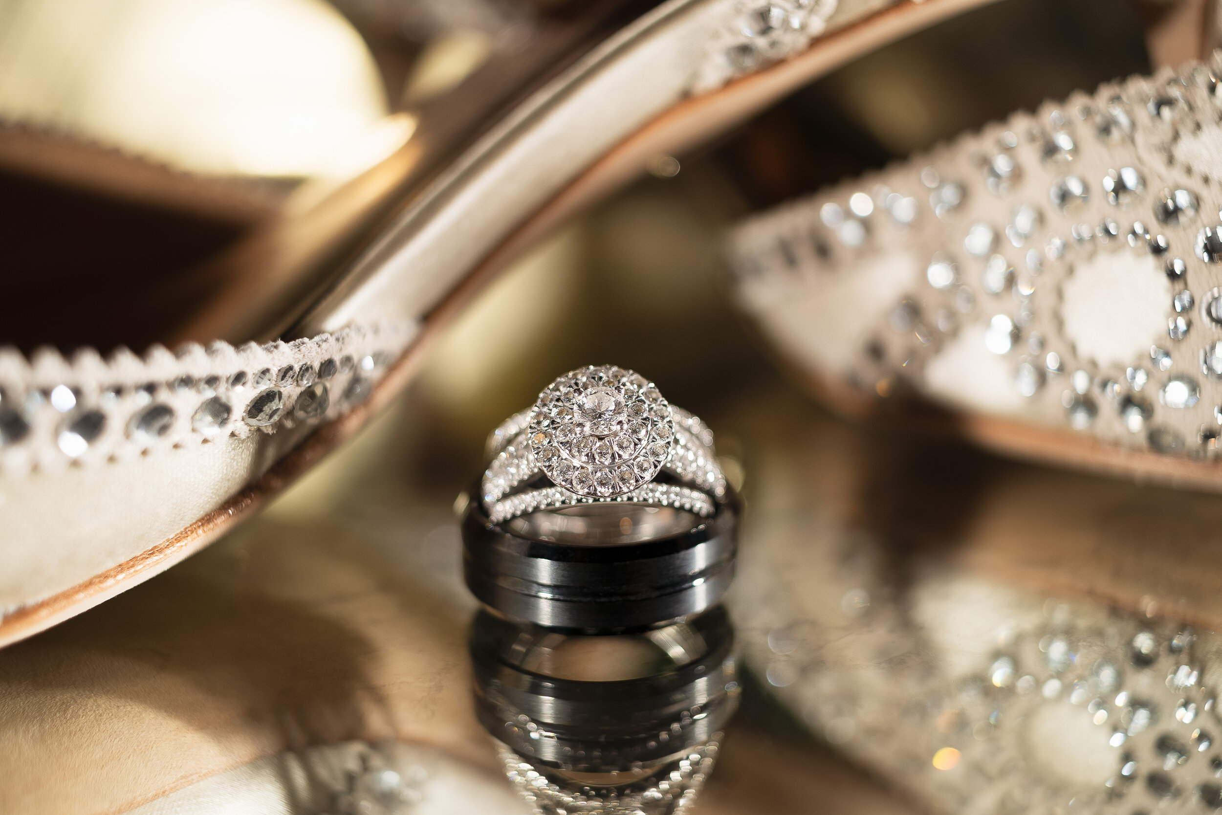 Close-up of a diamond engagement ring and wedding band on a reflective surface, with rhinestone-decorated wedding shoes in the background.