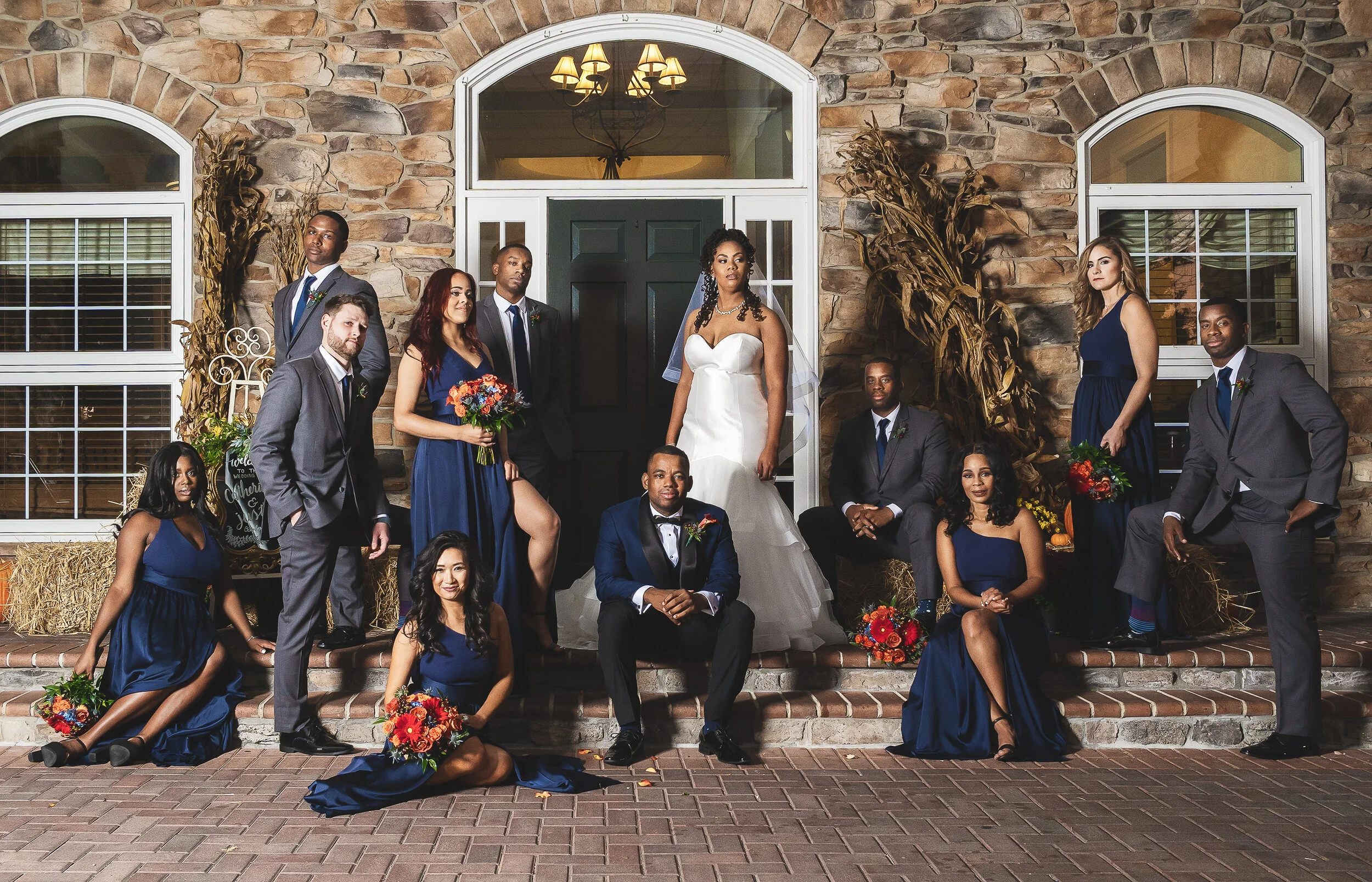 Wedding party posed in front of stone building with bride, groom, bridesmaids in blue dresses, and groomsmen in suits.