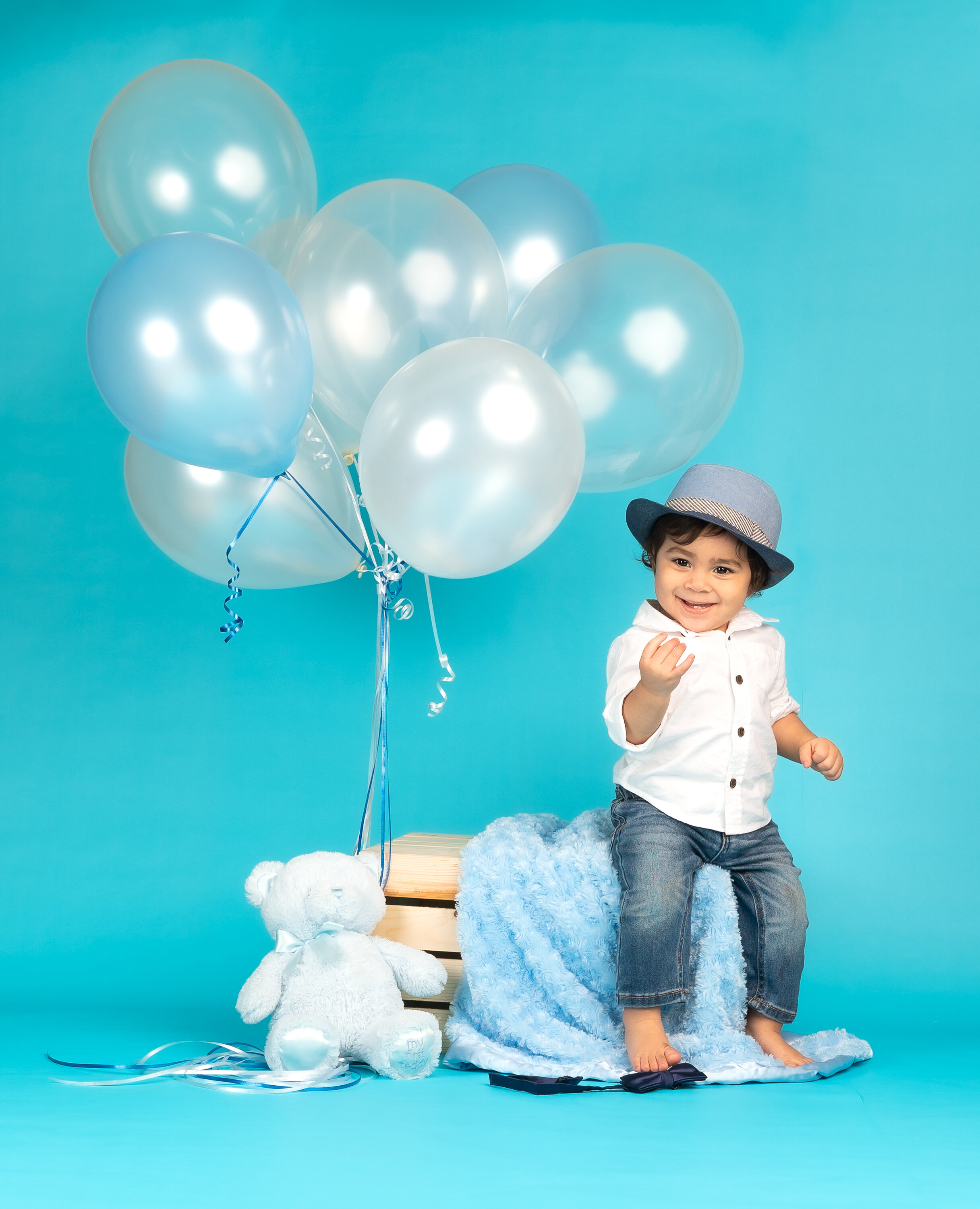 Smiling toddler in jeans and white shirt with a blue hat, sitting on a crate with light blue balloons and a teddy bear in the background, on a blue background.