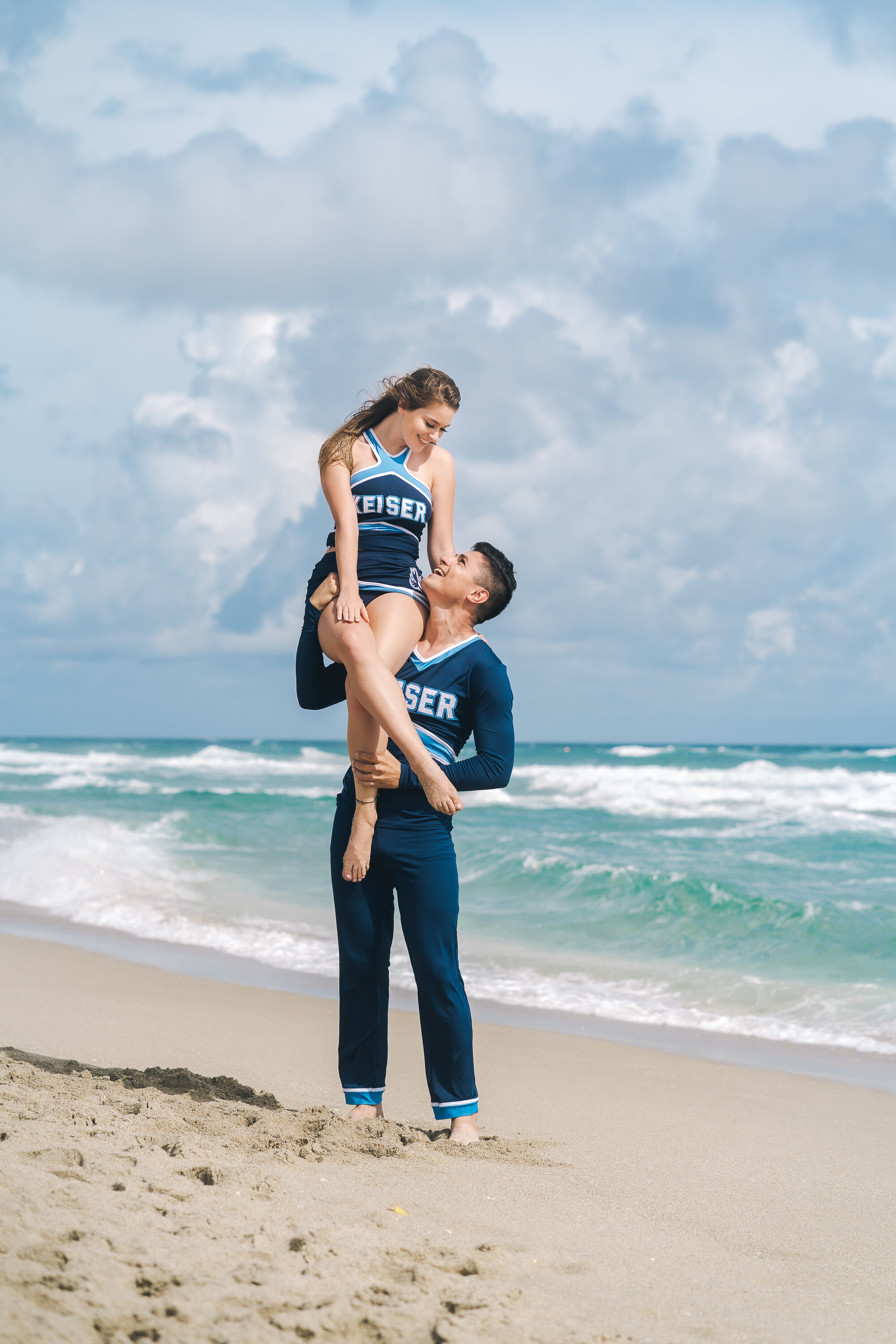 Two cheerleaders in blue uniforms, one male and one female, practicing a lift on a sandy beach with the ocean in the background.