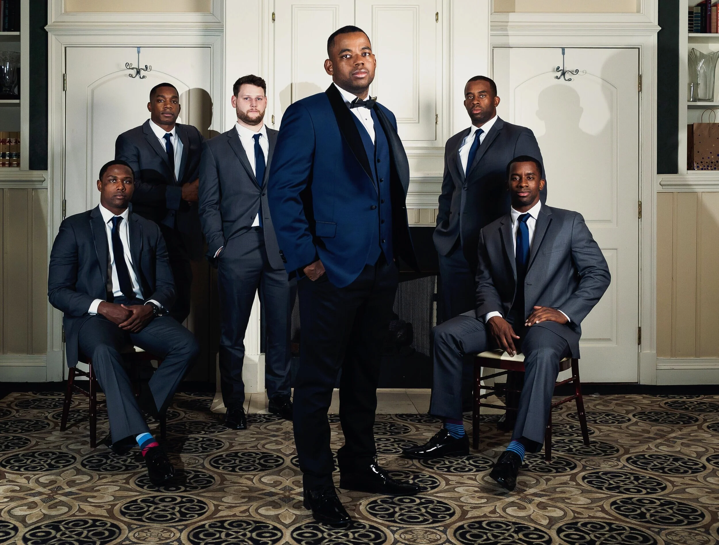 A group of six men in formal suits pose in an elegant indoor setting. Two are seated, while four stand. The room features patterned carpet and classic cabinetry.