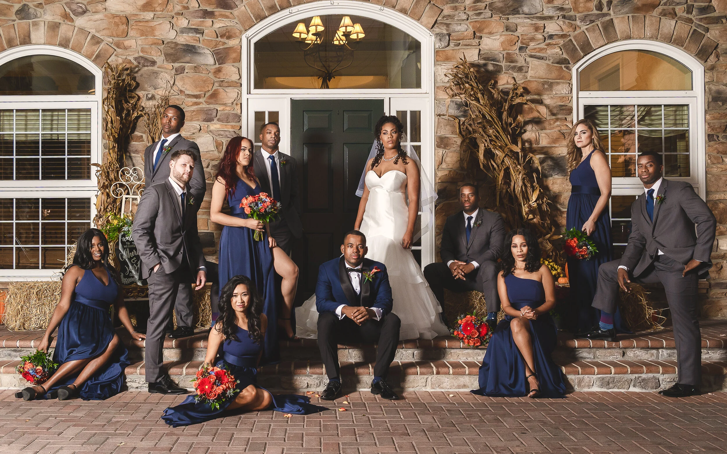 Bridal party posing in formal attire in front of a stone building, with bridesmaids in blue dresses and groomsmen in gray suits.