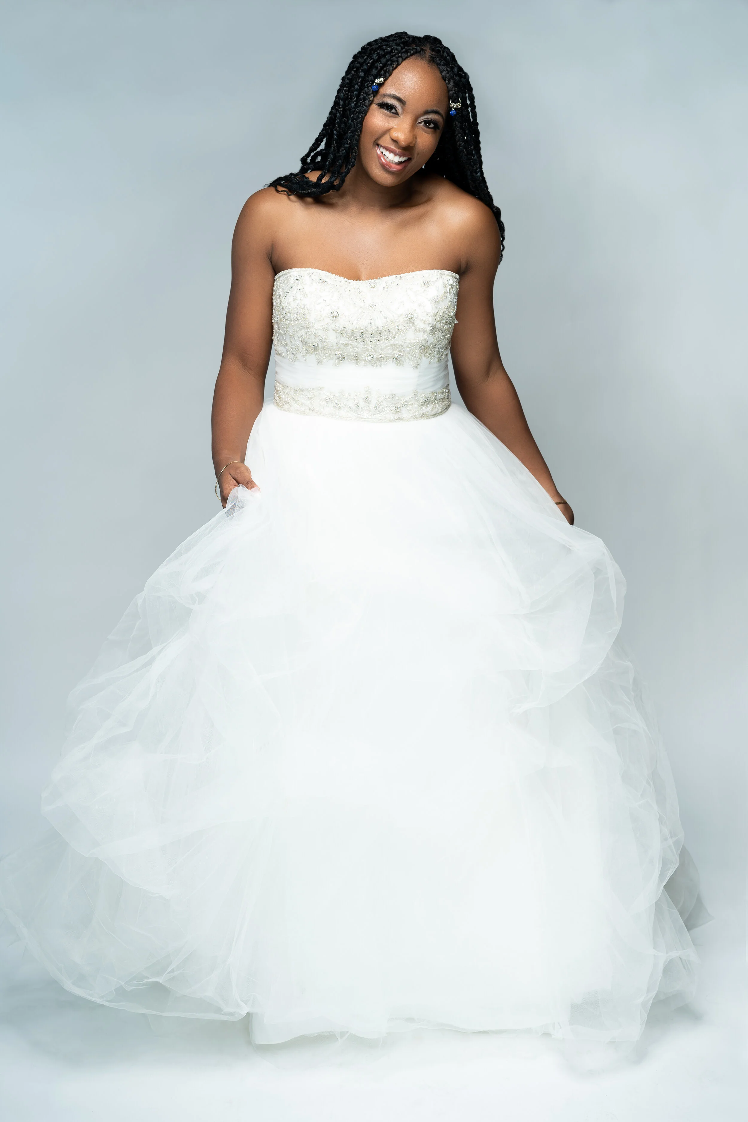 Smiling person in a strapless white wedding gown with intricate embroidery, holding the skirt, standing against a light gray background.