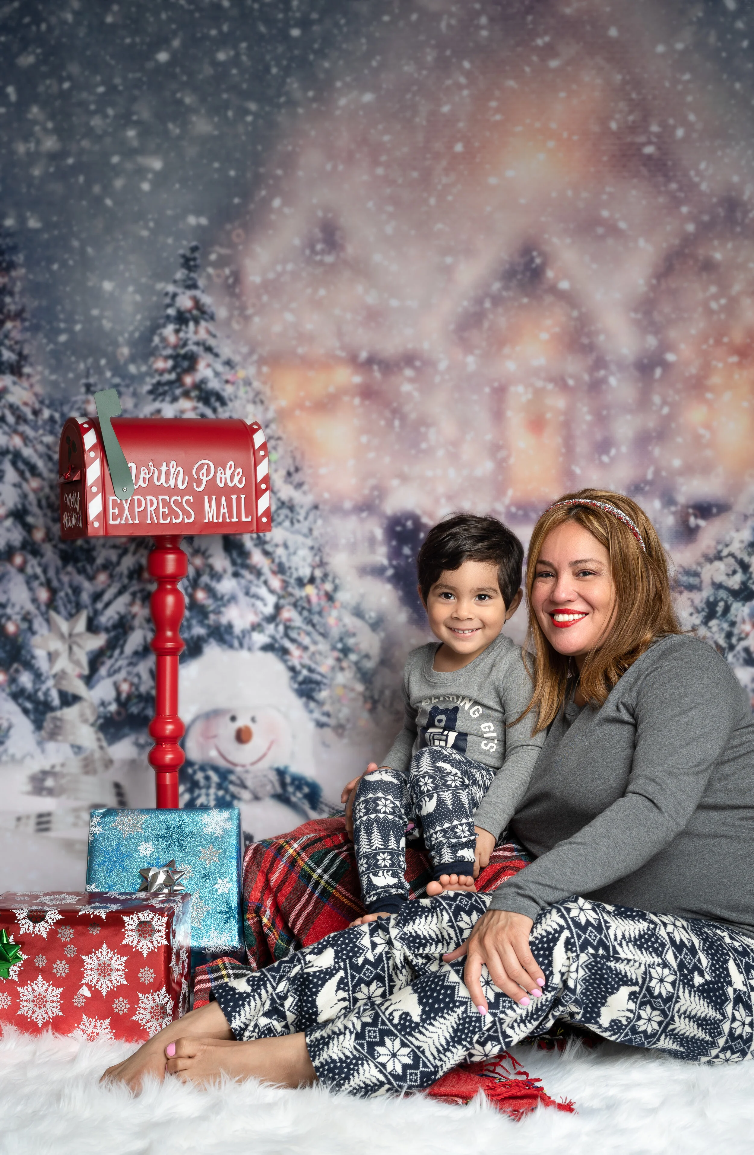 Mother and child in matching pajamas sitting by a decorated Christmas scene with snow, a North Pole Express Mail mailbox, and wrapped gifts.
