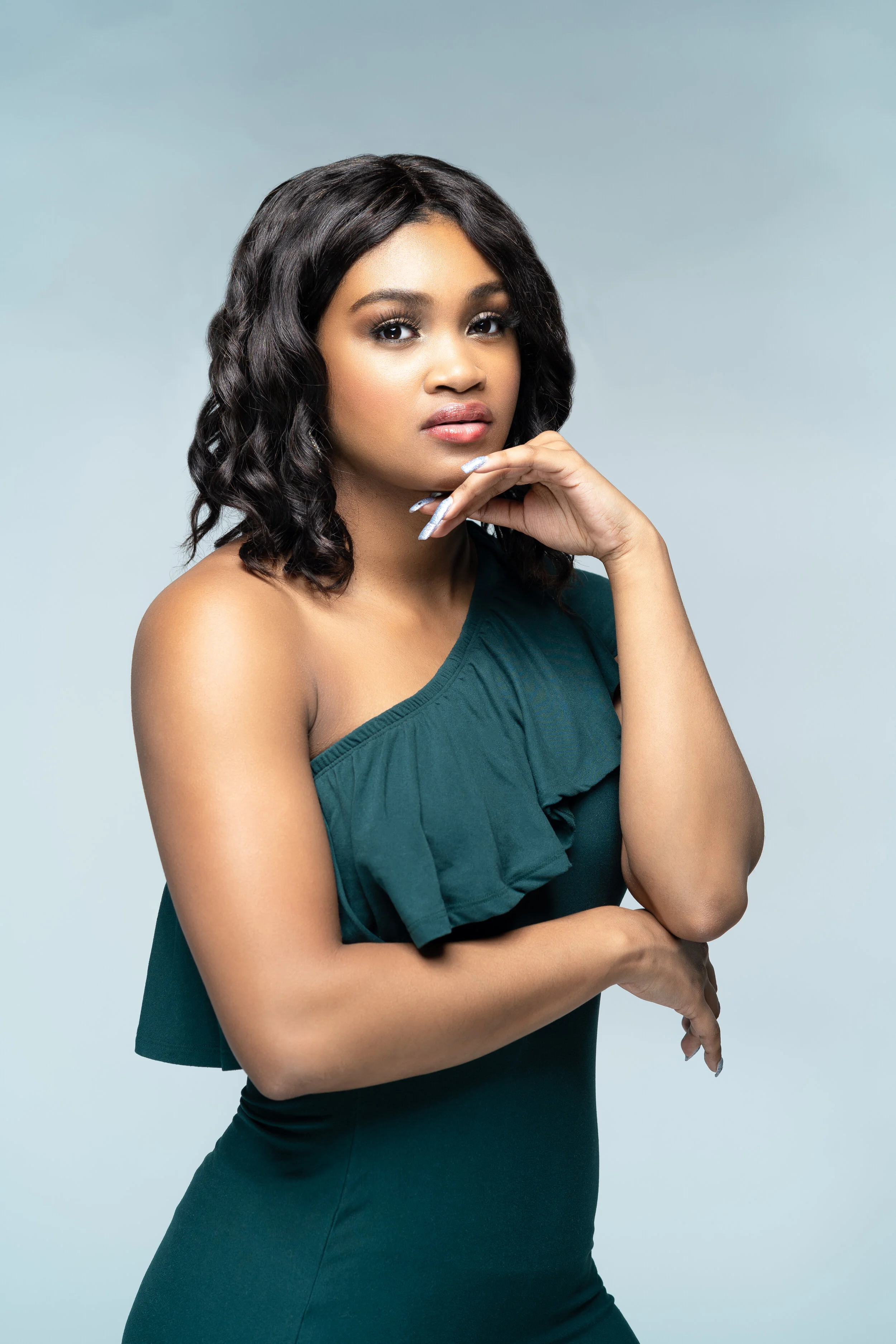 Woman in a green one-shoulder dress posing with hand on chin against a light gray background.