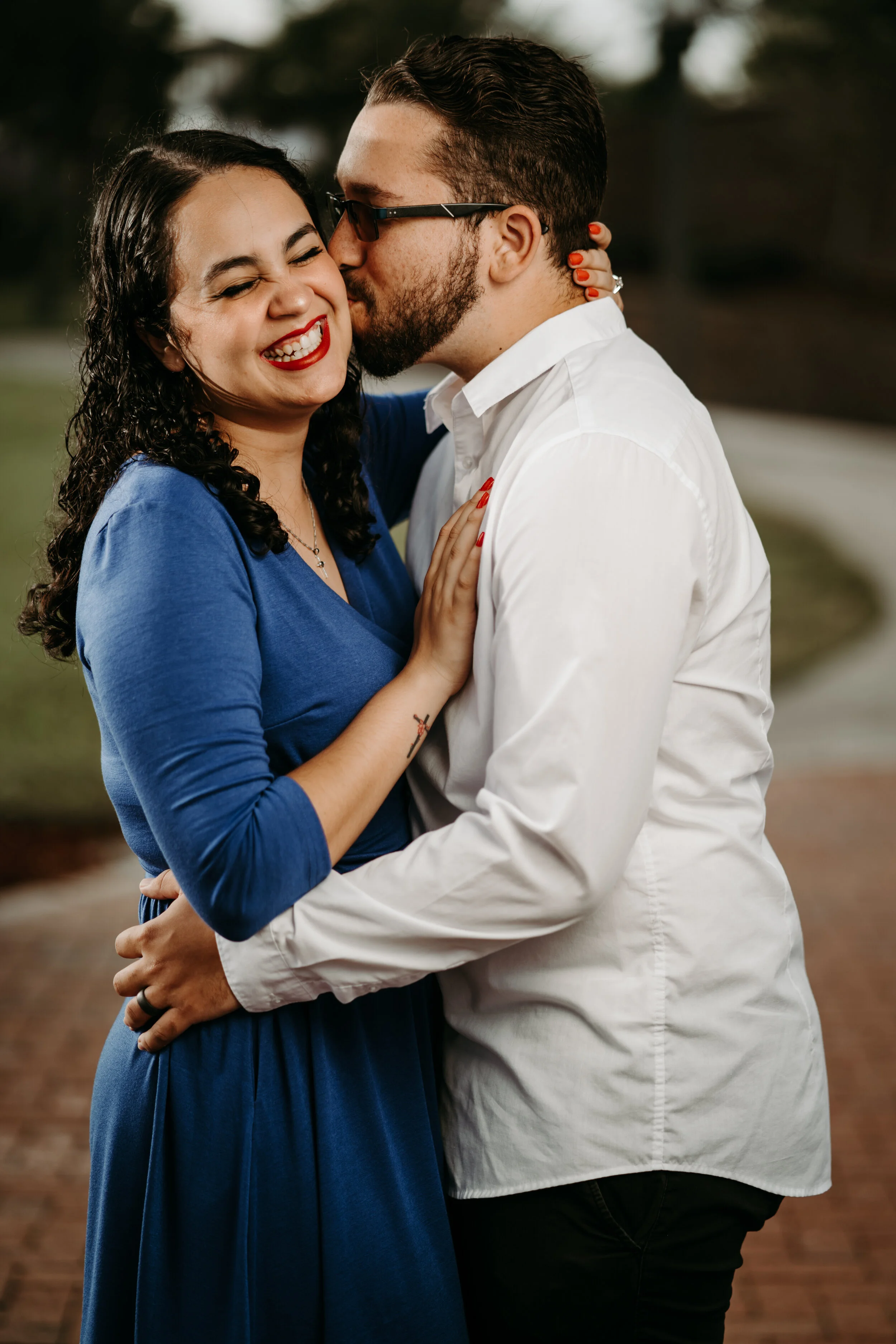 A couple embracing, with the man kissing the woman's cheek while she smiles, surrounded by an outdoor setting.