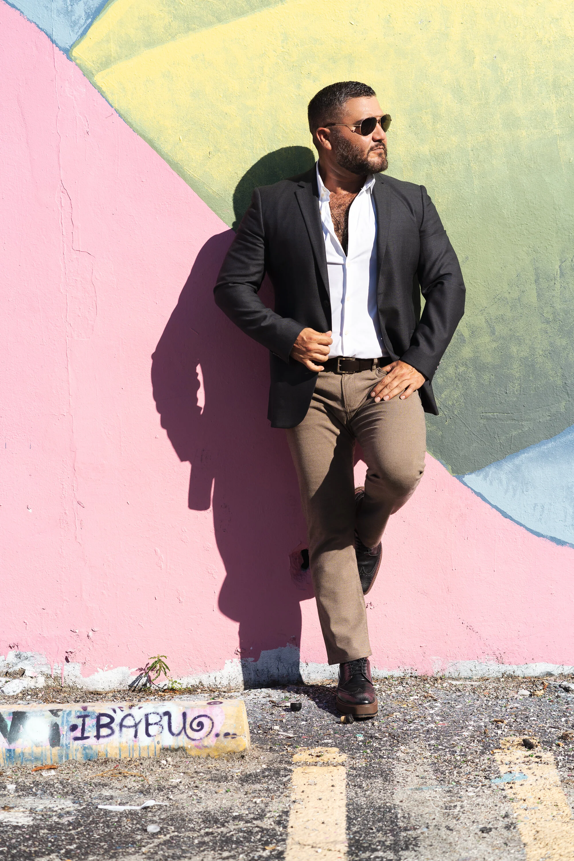 Man in sunglasses leaning against a colorful graffiti wall, wearing a black blazer, white shirt, and brown pants, standing in a parking lot.