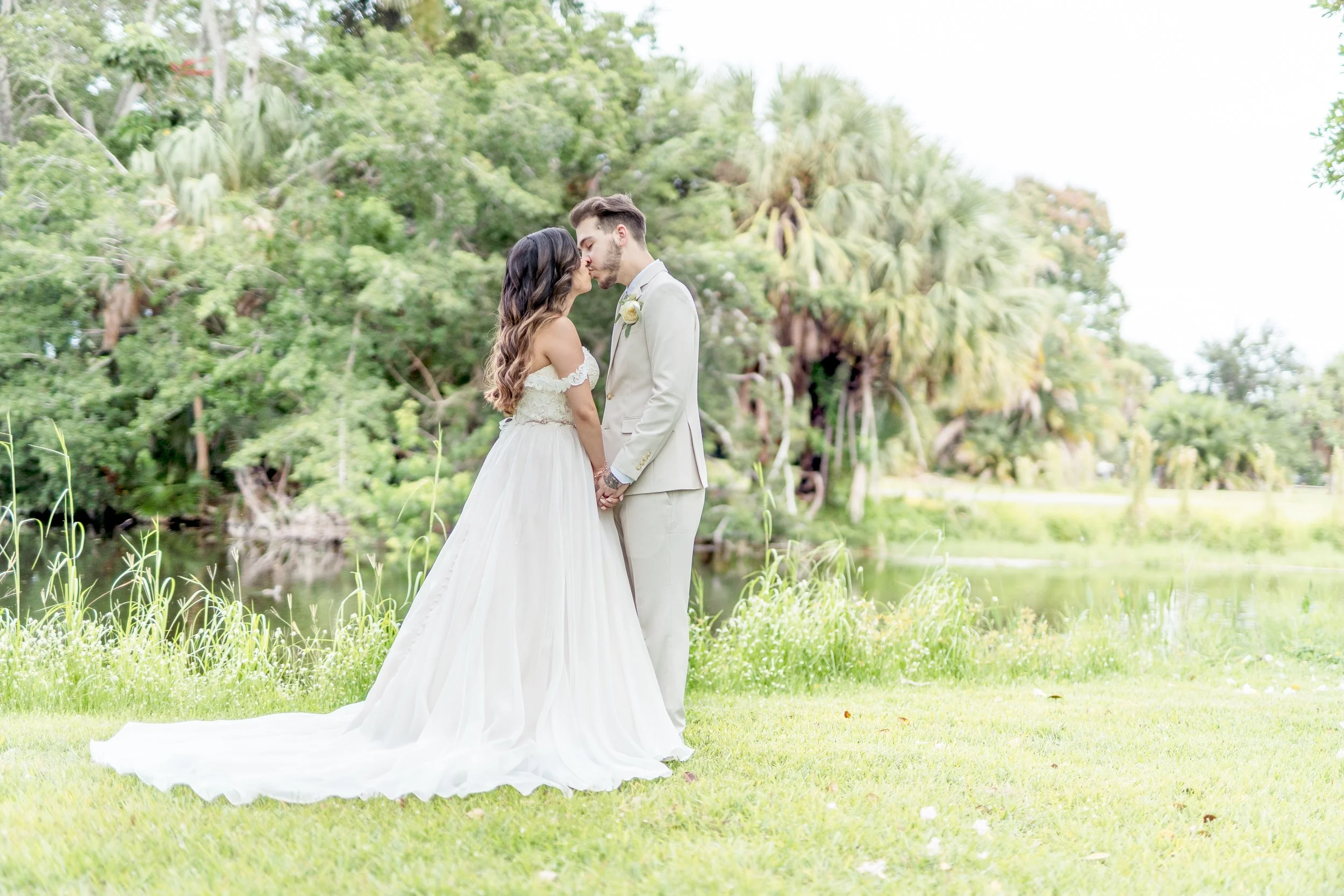 Bride and groom kissing outdoors next to trees and a pond.