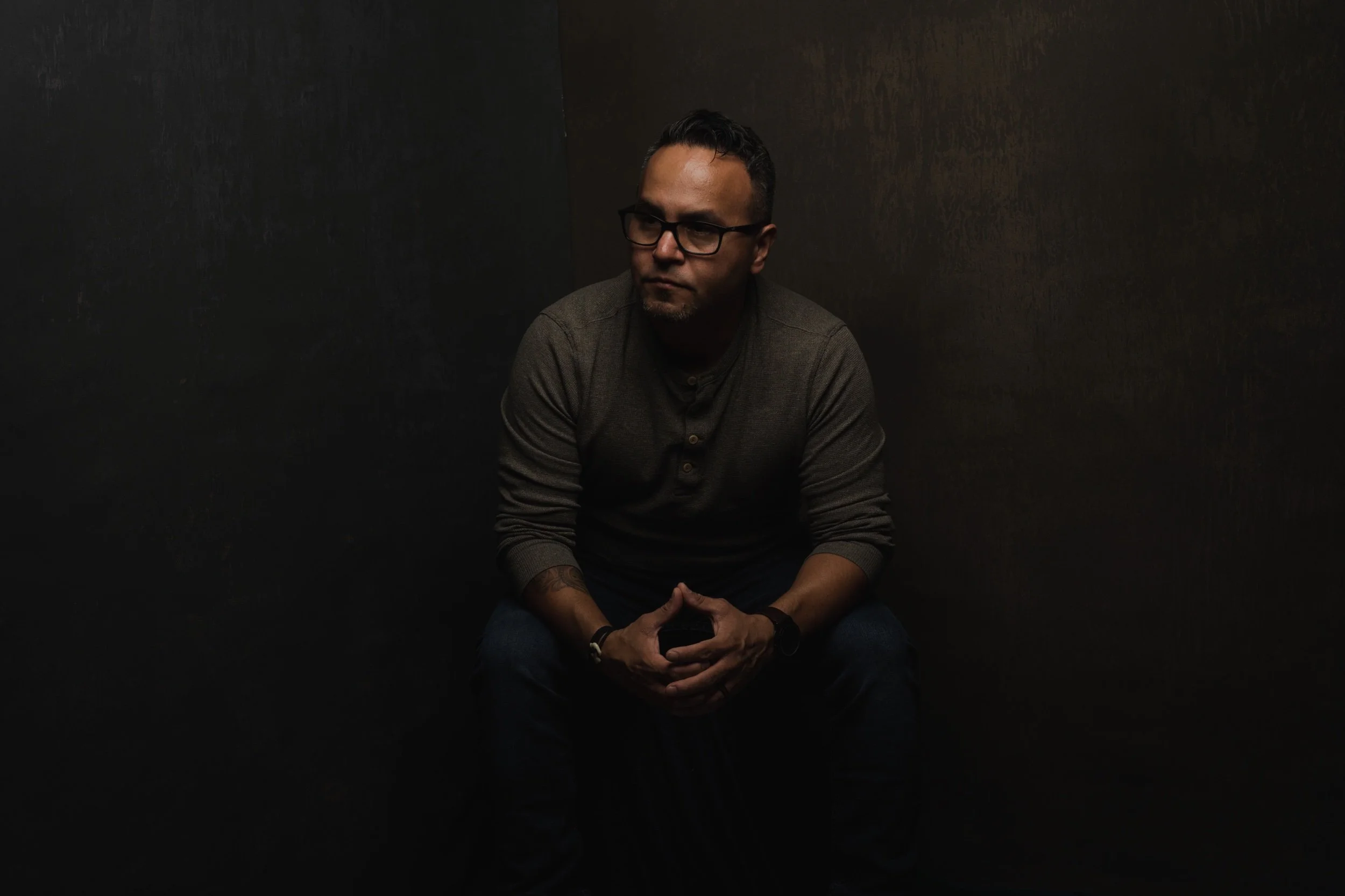 Man wearing glasses and a gray shirt sitting against a dark background, hands clasped.