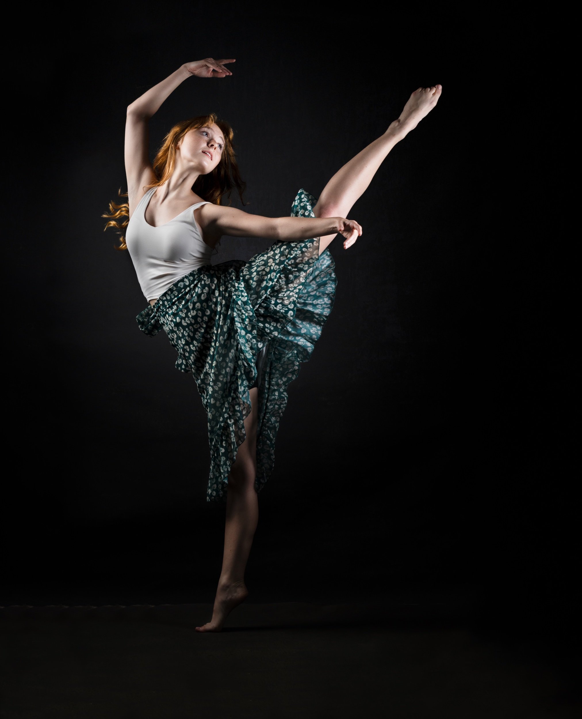 A dancer performing a high kick with one leg extended, wearing a white top and a patterned skirt, set against a dark background.
