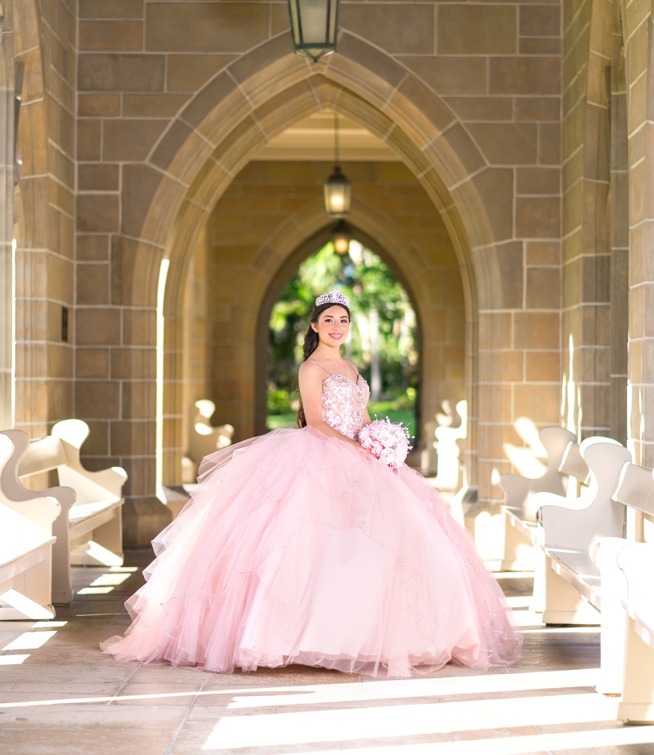 Young woman in a pink ball gown and tiara standing in an arched hallway, holding a bouquet, with sunlight streaming through.