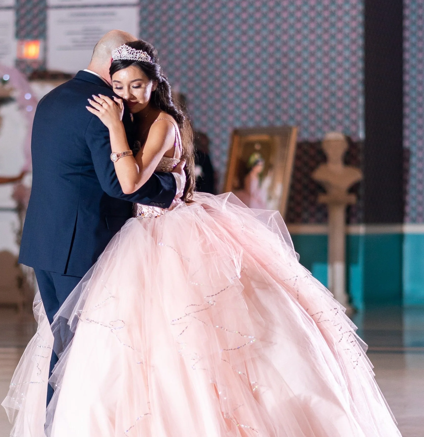 A young woman in a pink ball gown and tiara dances with a man in a suit at an indoor event.