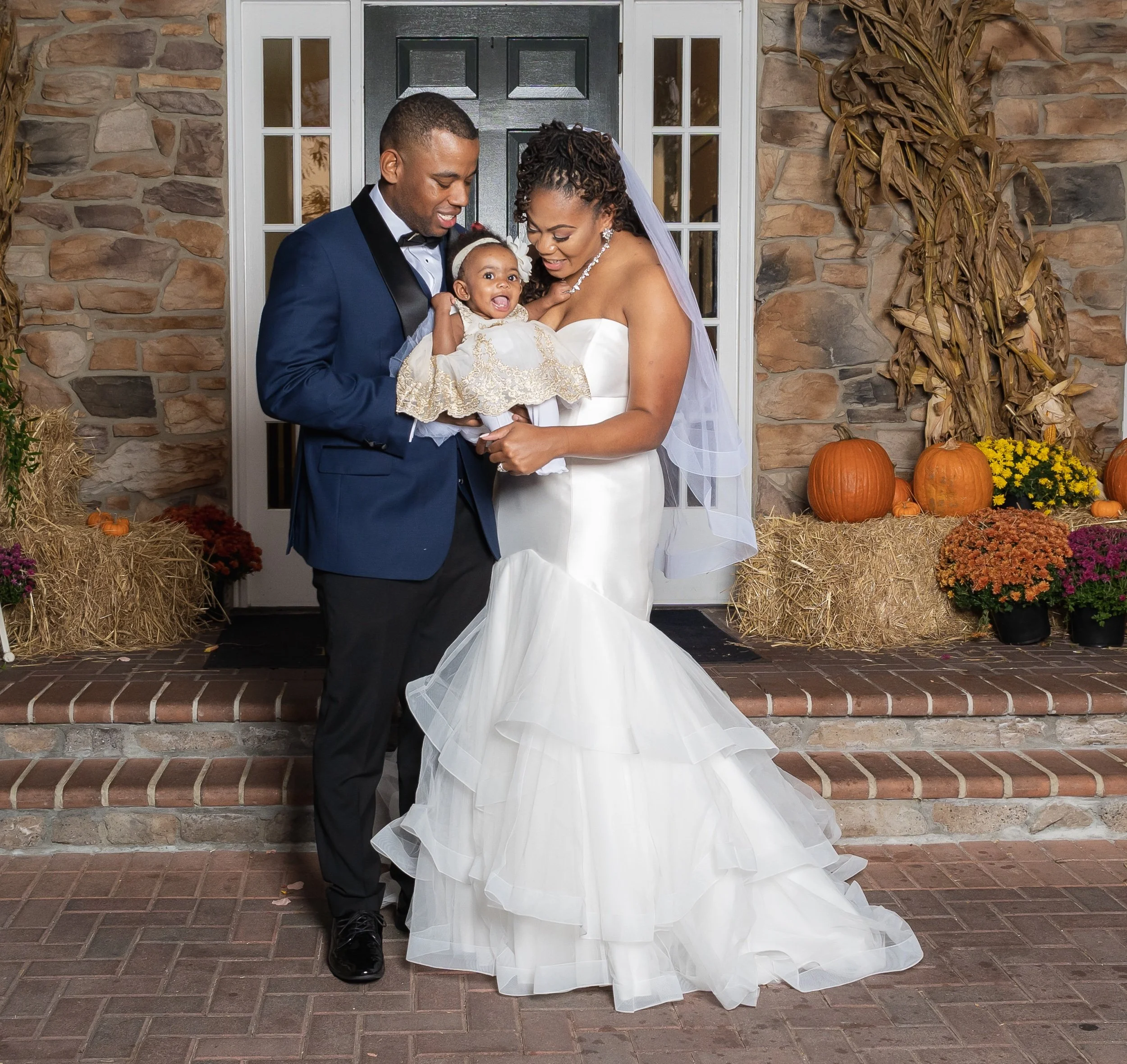 A couple dressed formally, with the woman in a white wedding gown and the man in a dark suit, holding a smiling baby wearing a white dress, in front of a decorated entrance with pumpkins and hay.