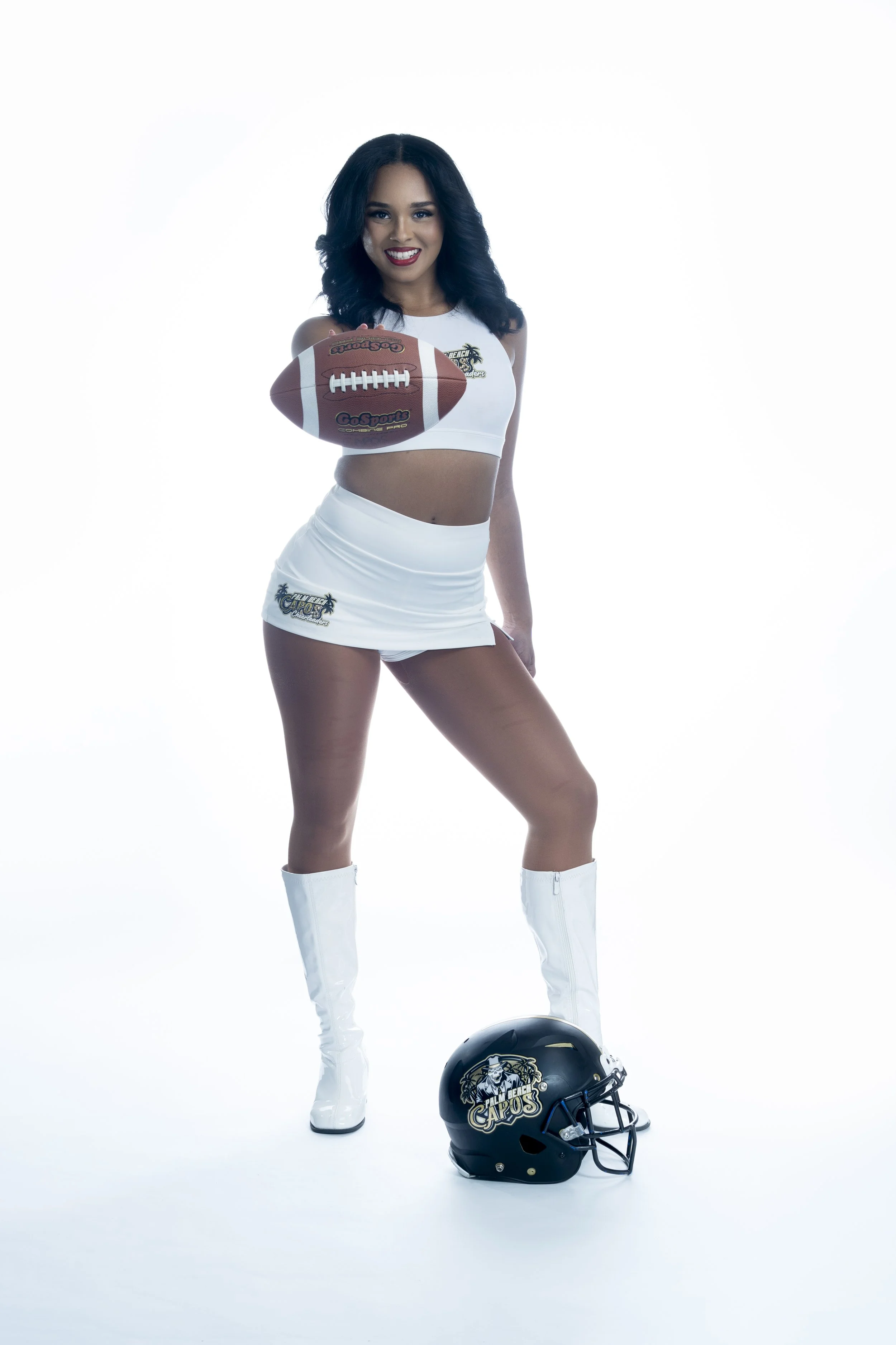 Woman in cheerleading outfit holding a football, wearing white boots, with a football helmet on the ground.