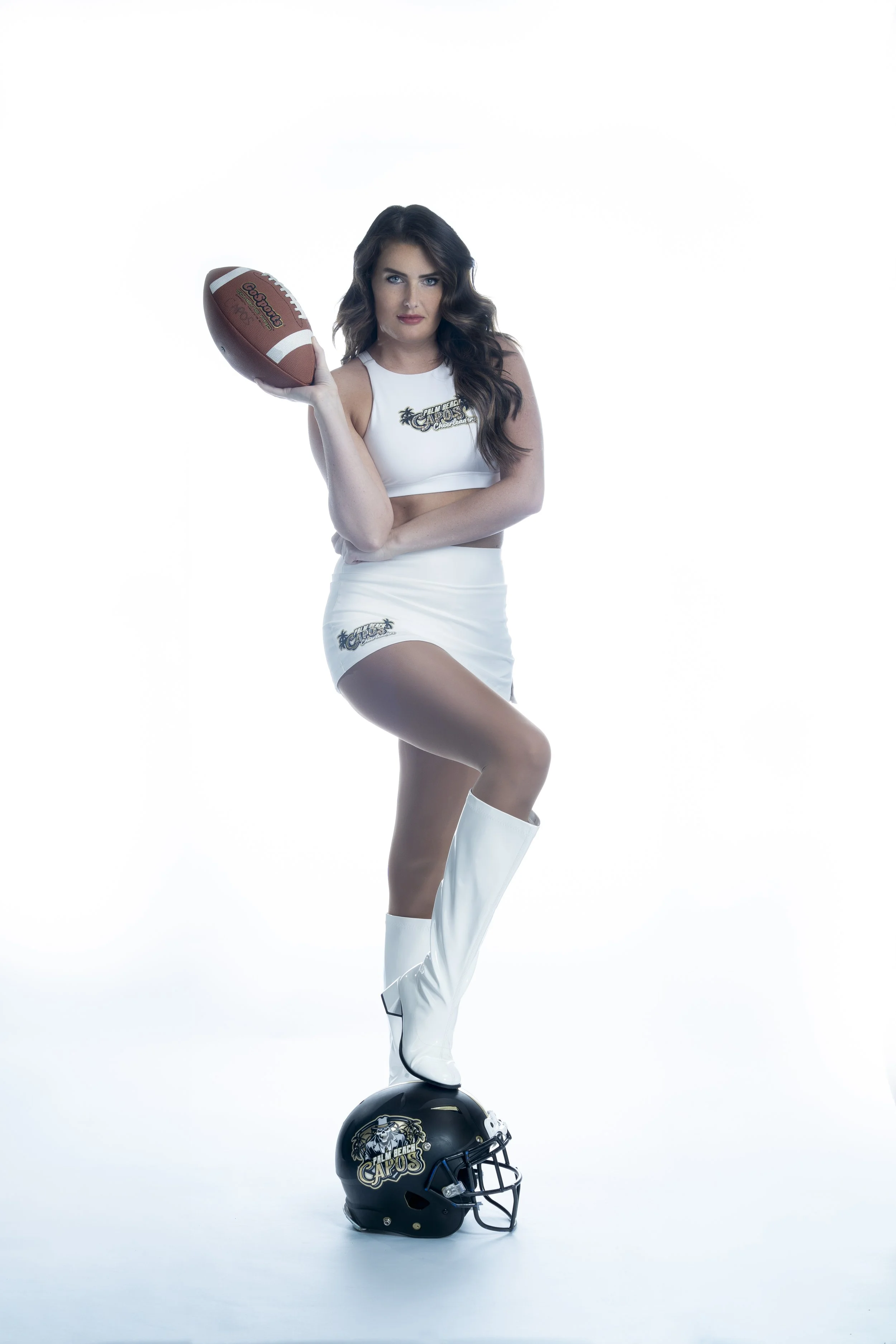Woman in white outfit holding a football, standing with one leg on a helmet.