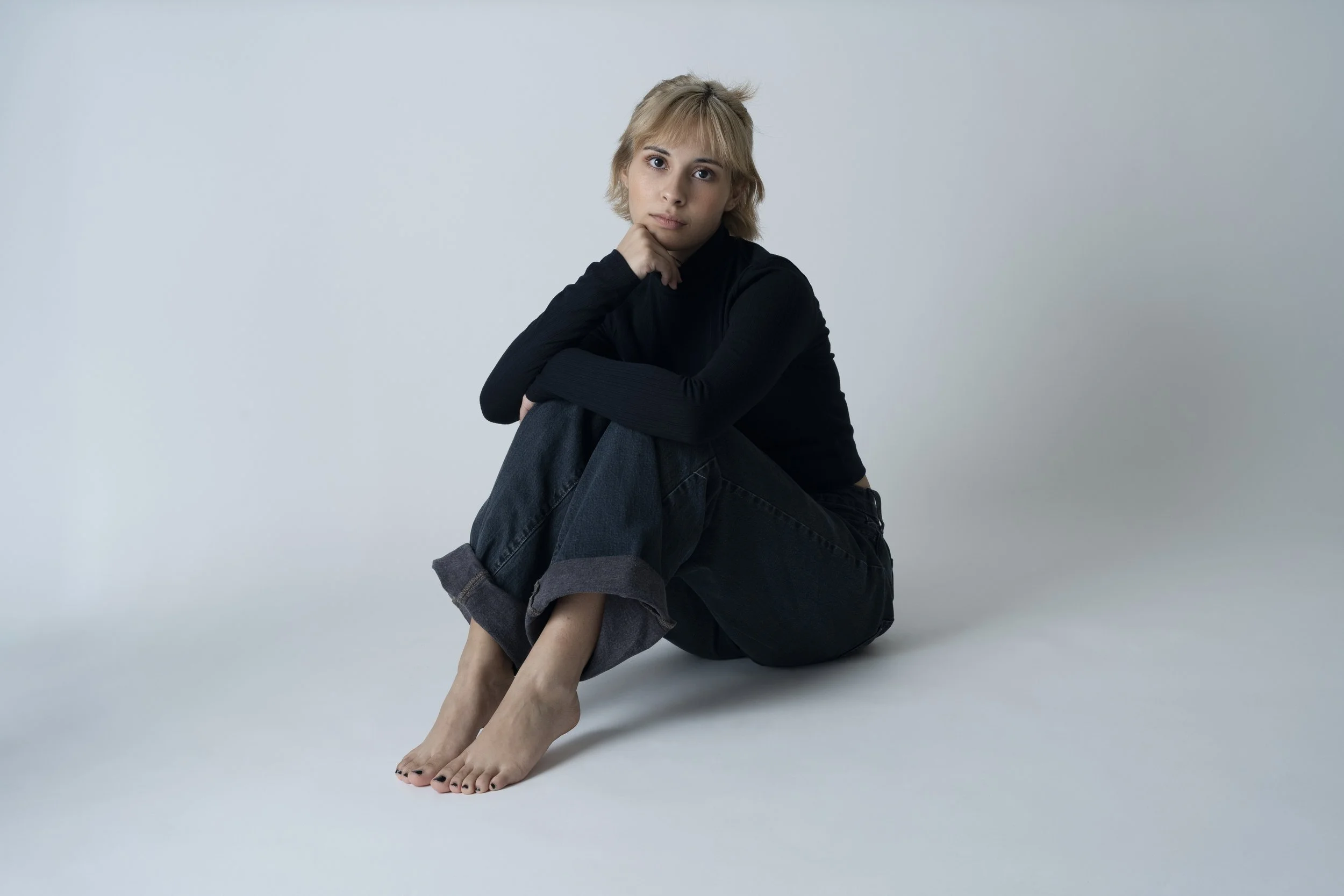 Person sitting on the floor with knees up, wearing a black long-sleeve shirt and jeans, with a thoughtful expression.