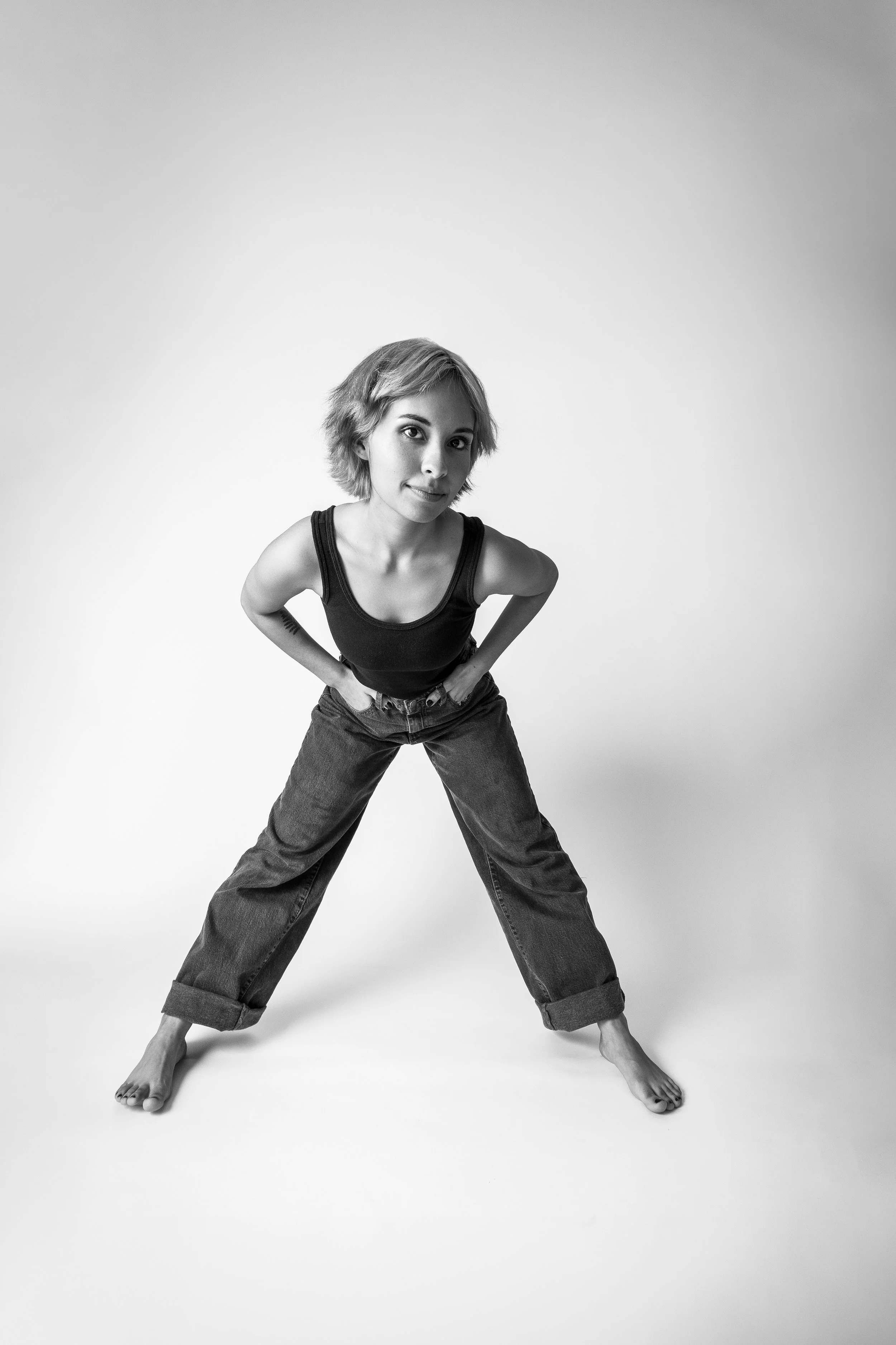 Black and white photo of a person posing with feet apart, hands on hips, wearing a tank top and jeans, against a plain background.
