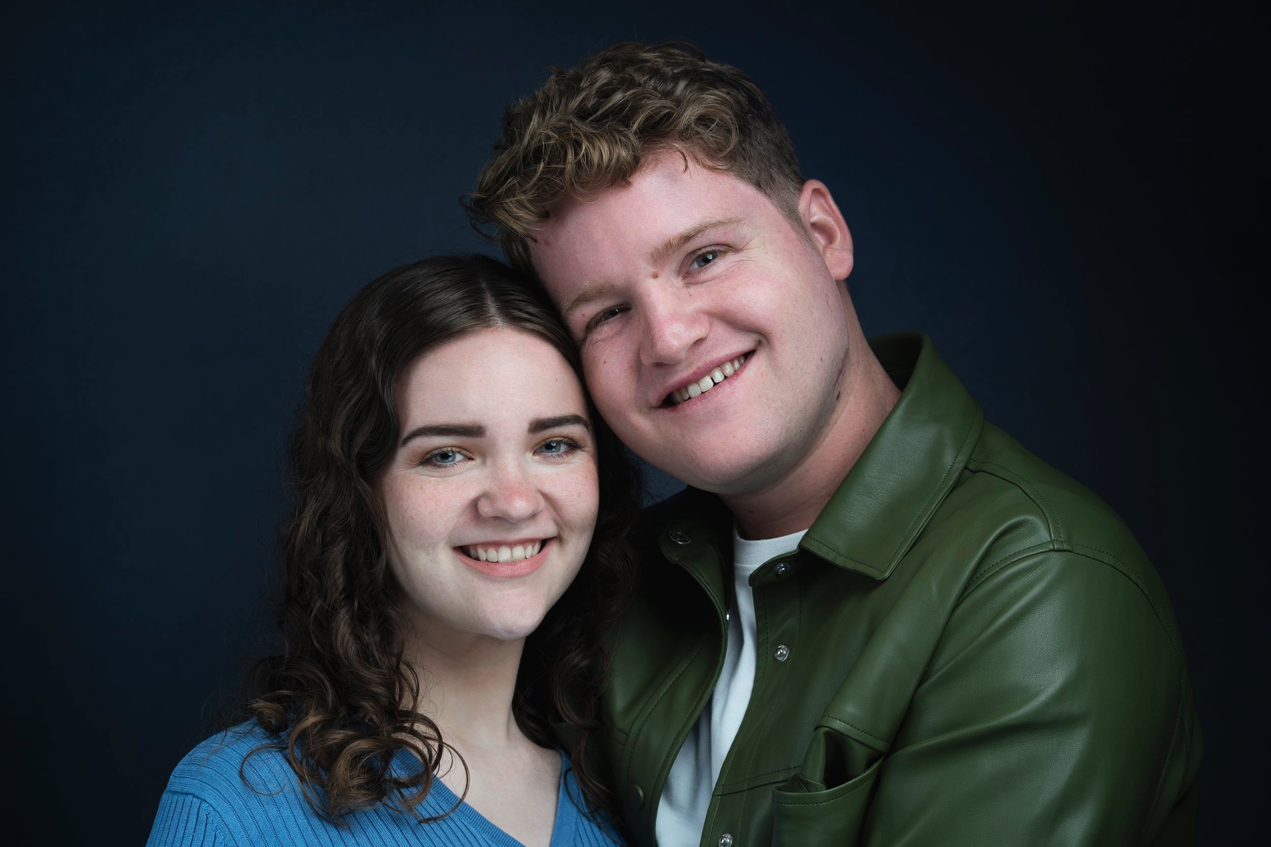 A smiling man and woman pose together against a dark background. The man wears a green jacket, and the woman wears a blue top.