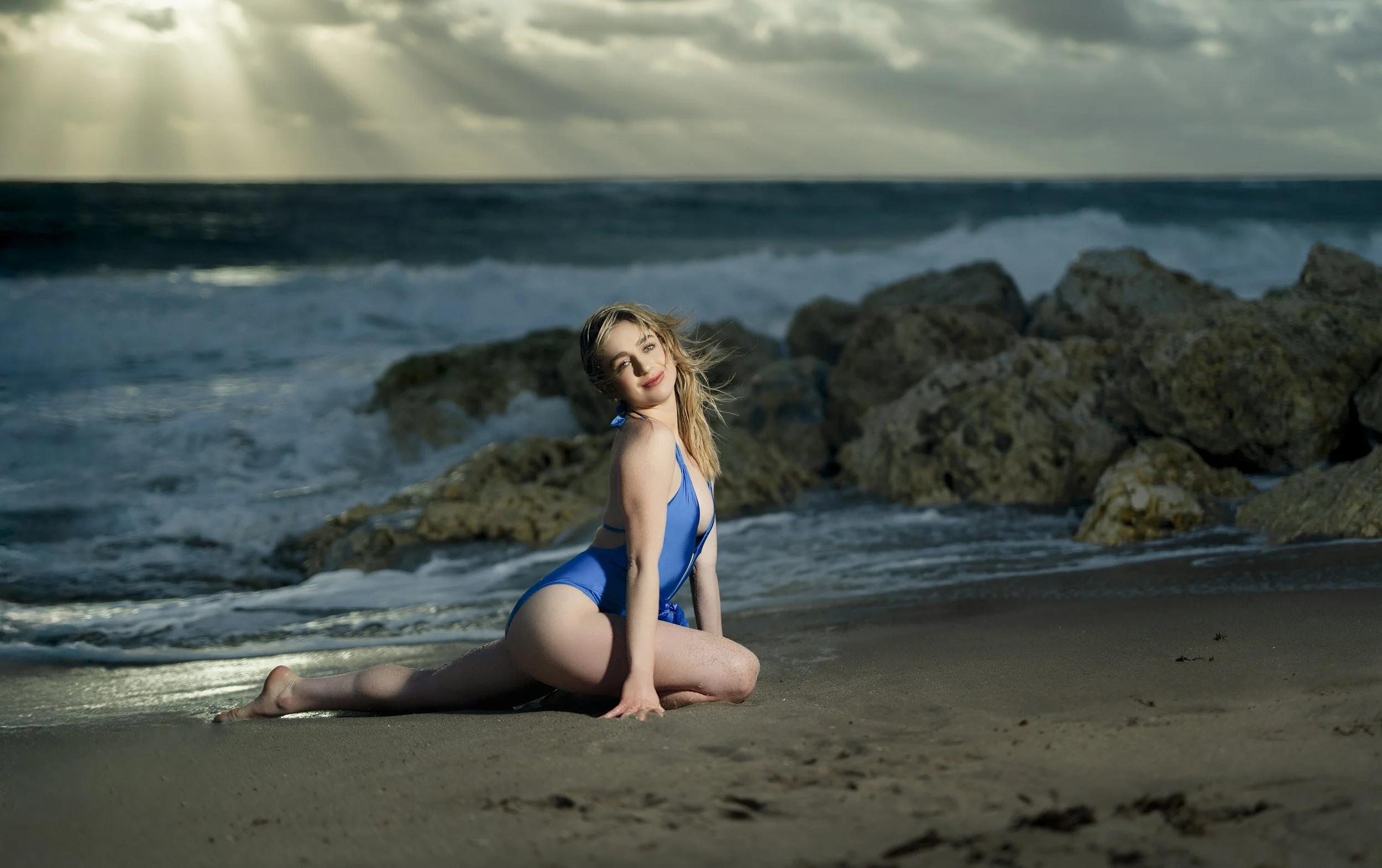 A woman in a blue swimsuit kneeling on the beach with rocks and ocean waves in the background, under cloudy and partly sunny skies in Palm Beach, FL. 