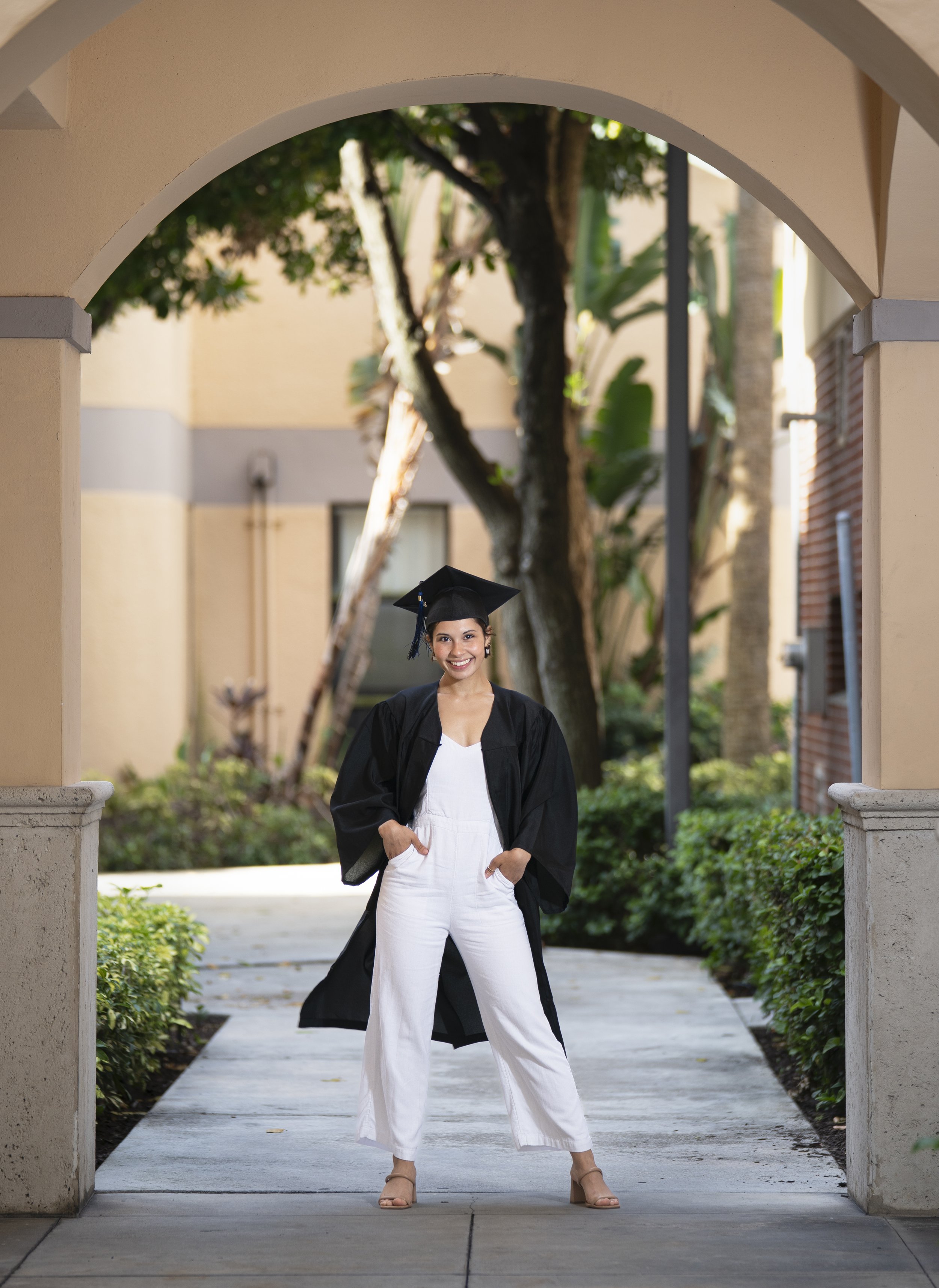 A woman in a black graduation gown and cap standing outdoors on a walkway, smiling with hands in pockets, with trees and a building in the background.