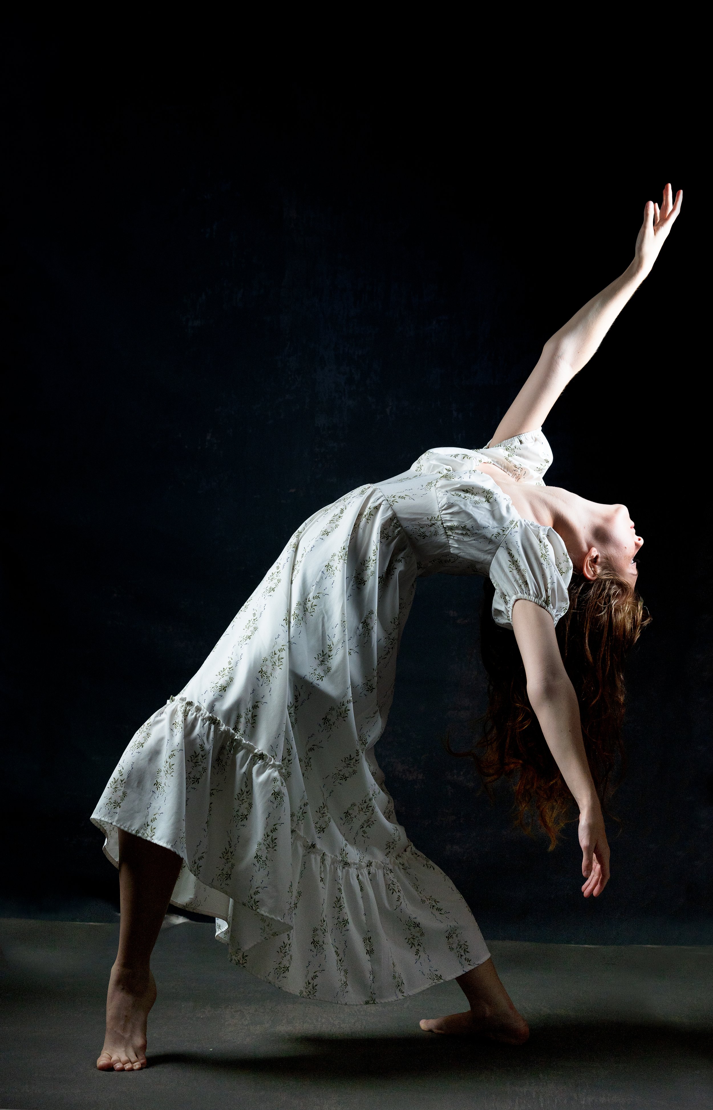 A woman in a white floral dress performs a graceful backbend dance pose against a dark background.