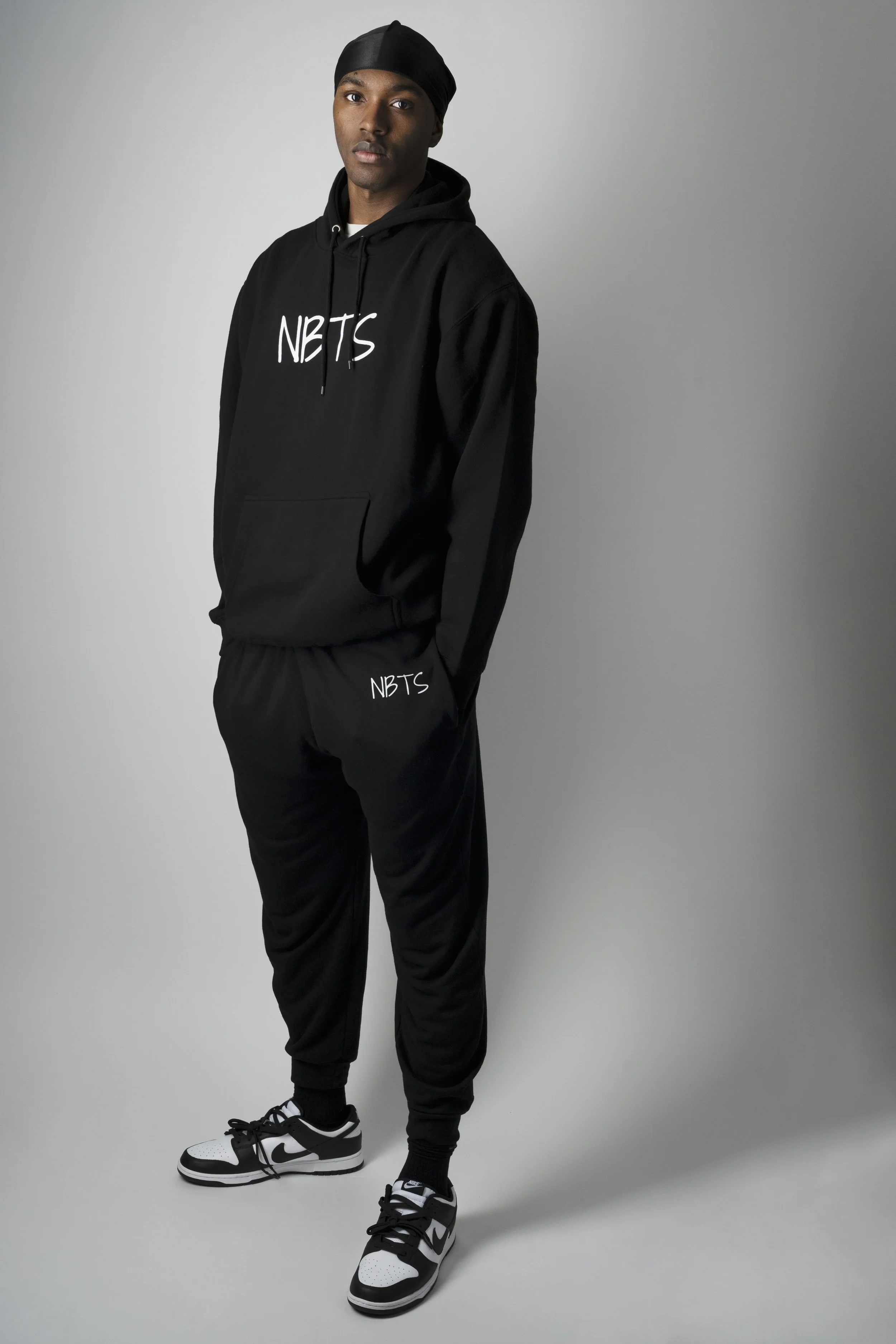 Person wearing a black "NBTS" hoodie and sweatpants, standing in a studio, with black and white sneakers.