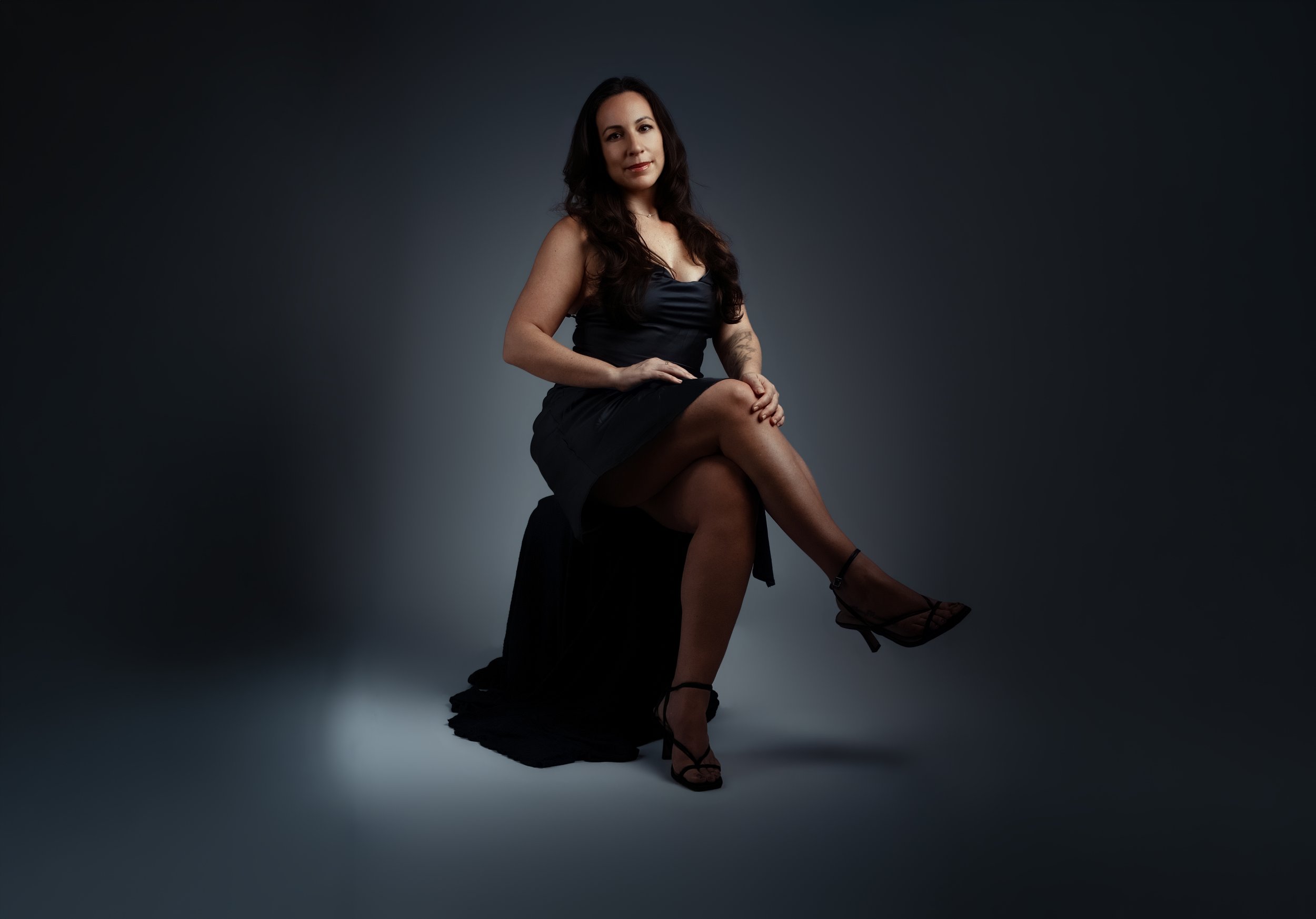 A woman sitting on a black box against a dark gradient background, wearing a black satin dress and high heels.