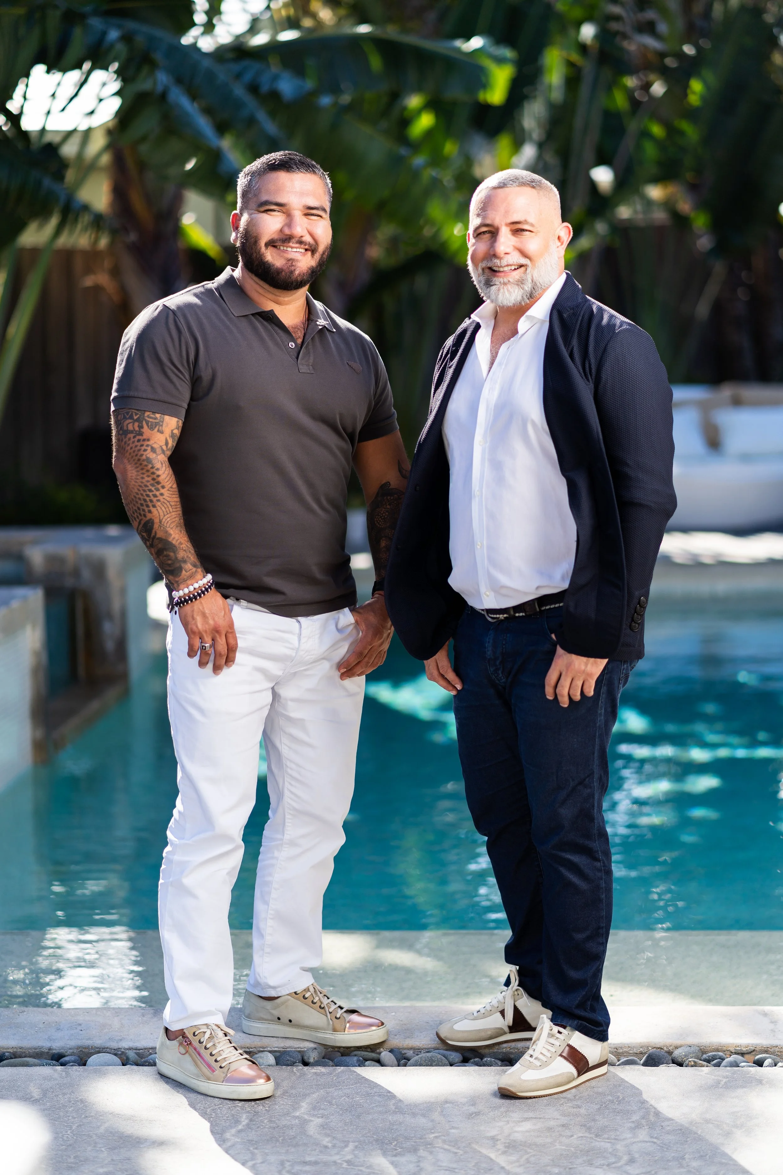 Two men smiling and standing by a swimming pool with tropical plants in the background.