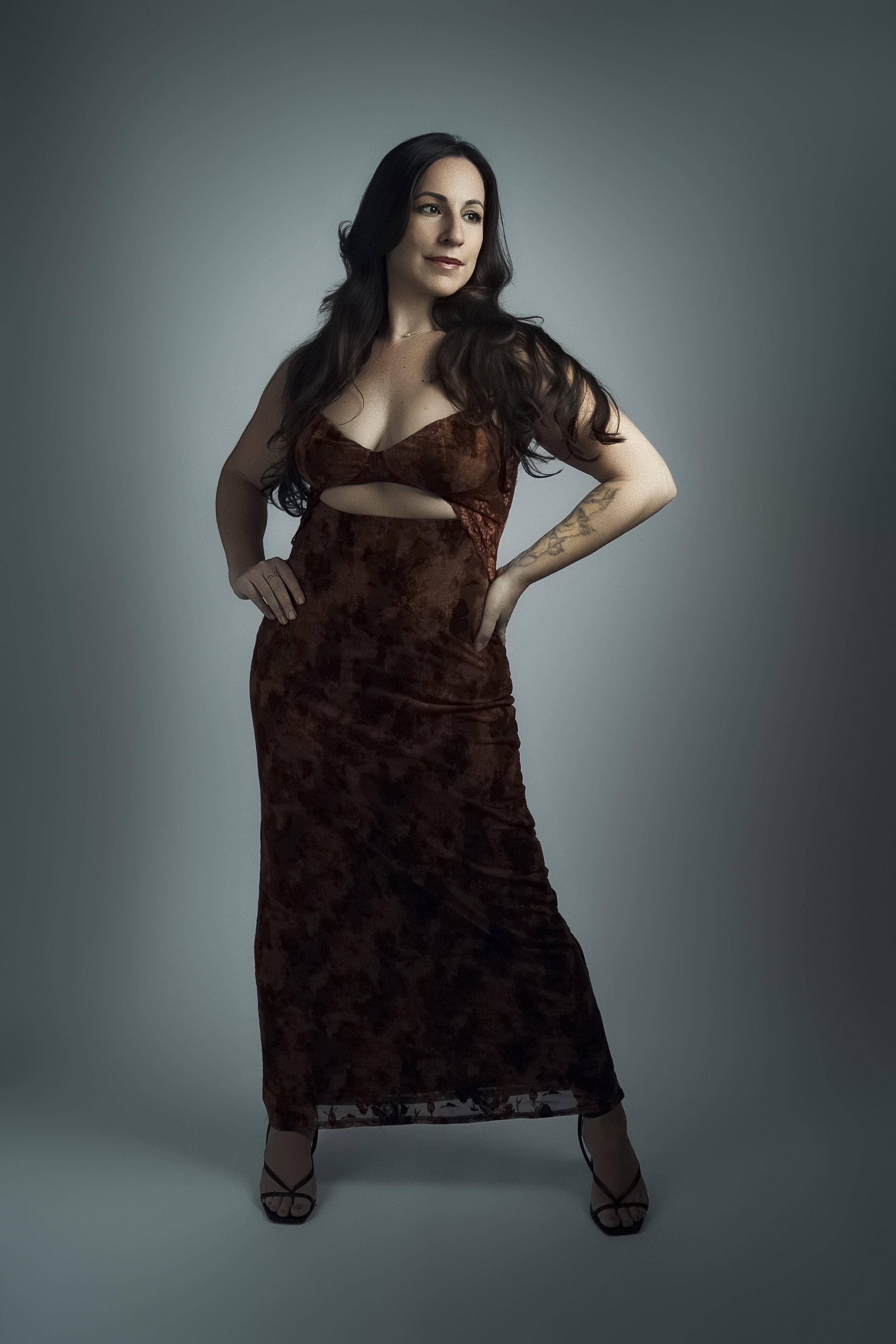 A woman with dark hair, wearing a brown velvet dress with a cutout, standing with one hand on her hip against a gradient gray background.