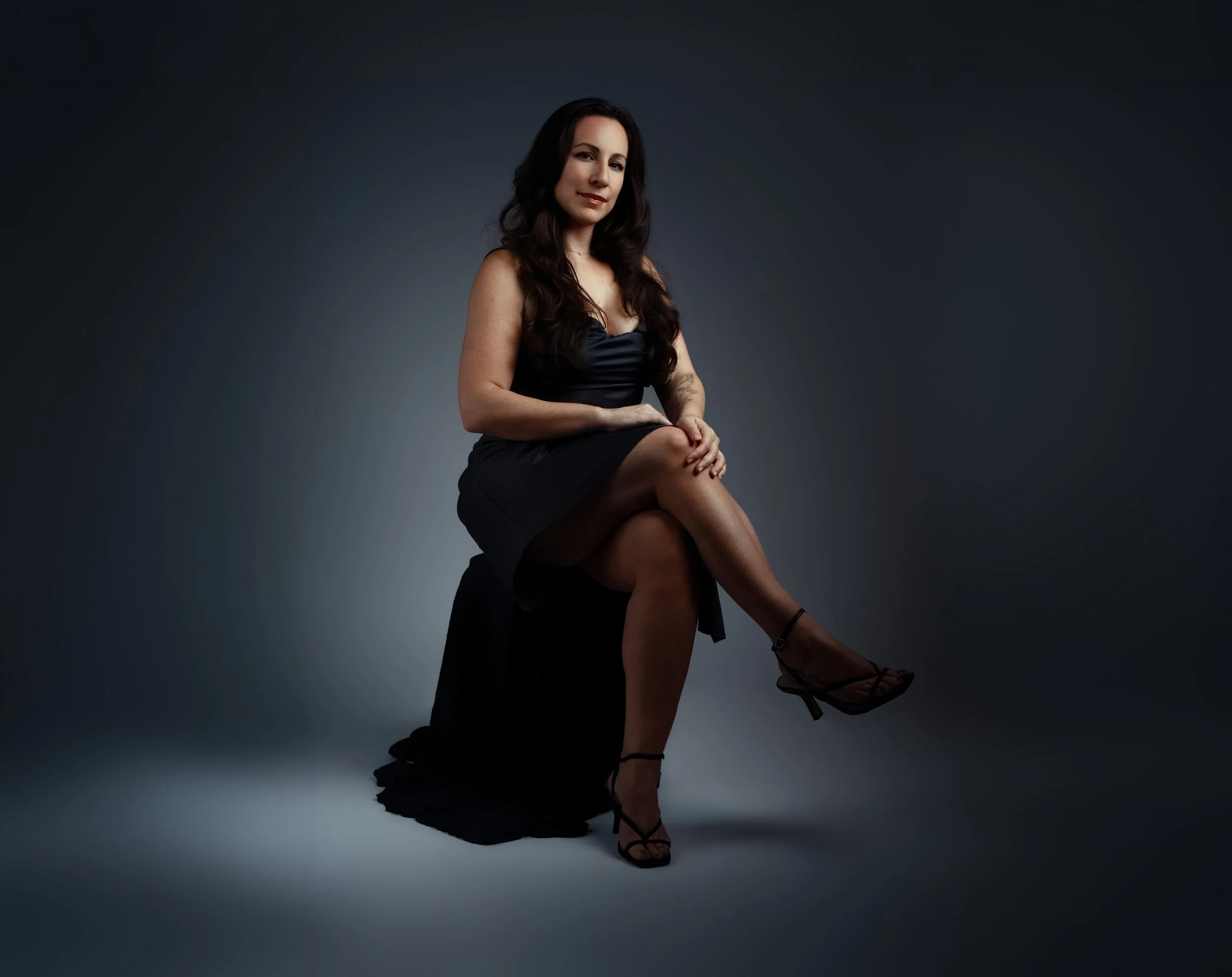 A woman sitting on the floor with her legs crossed, wearing a black dress and high heels, against a dark, neutral background.