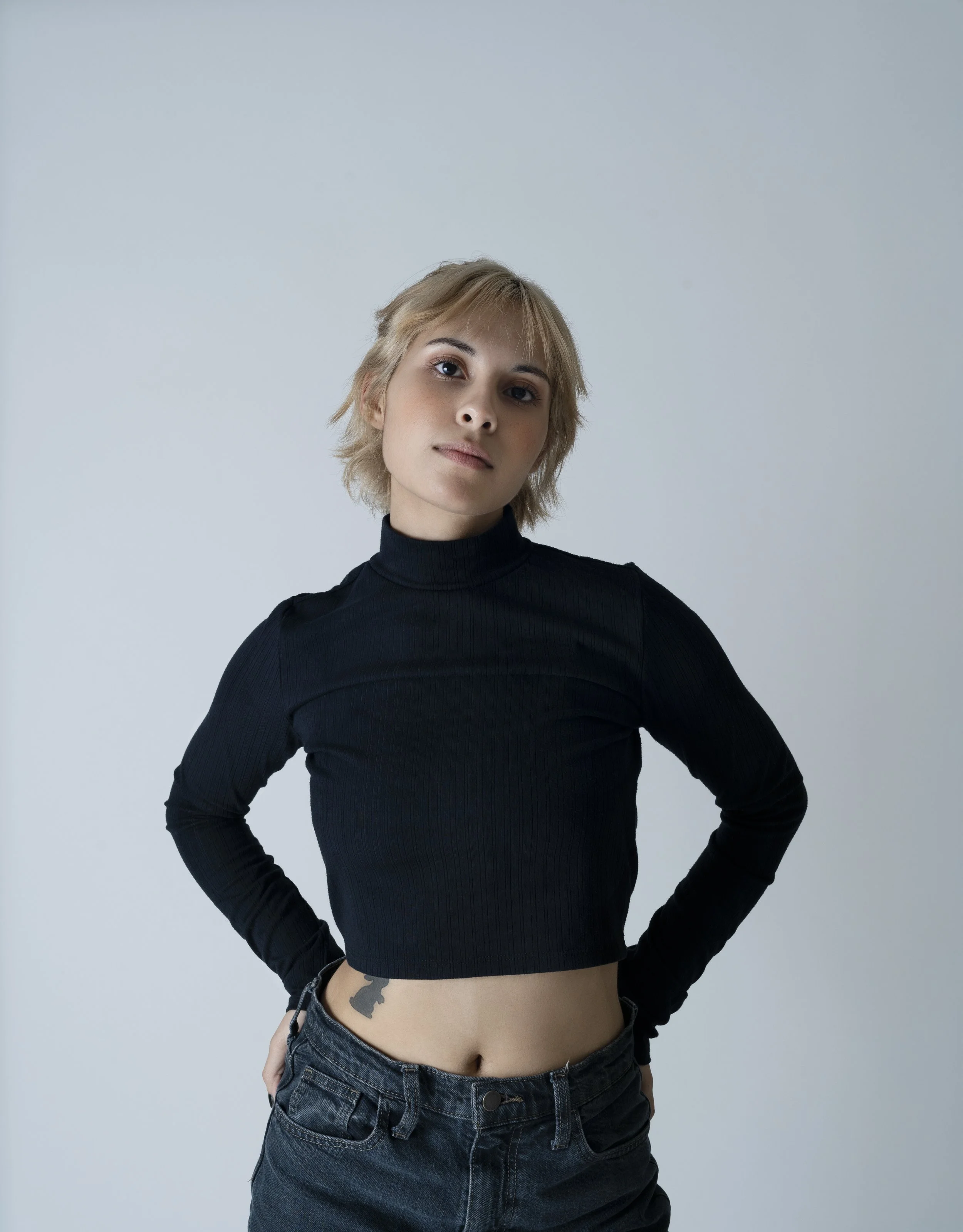 Person with short blond hair in black long-sleeve crop top and jeans against gray background.