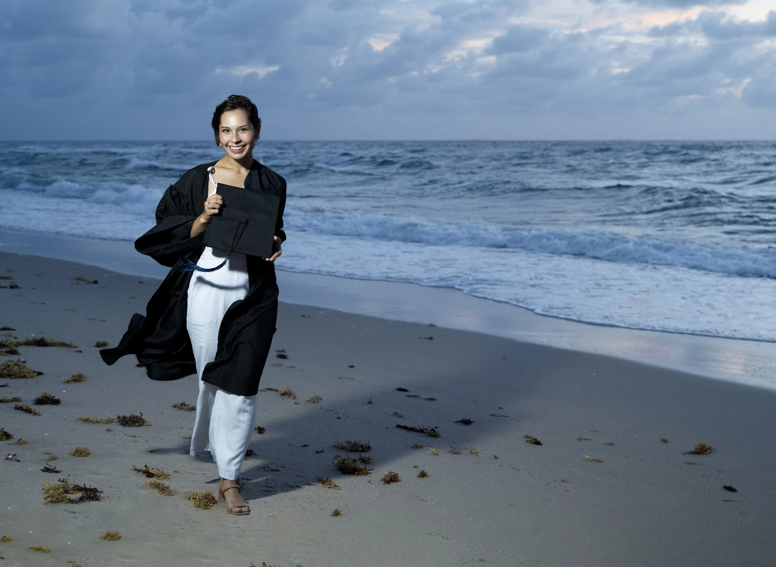 A woman in graduation cap and gown holding a diploma walking on the beach with the ocean and cloudy sky in the background.