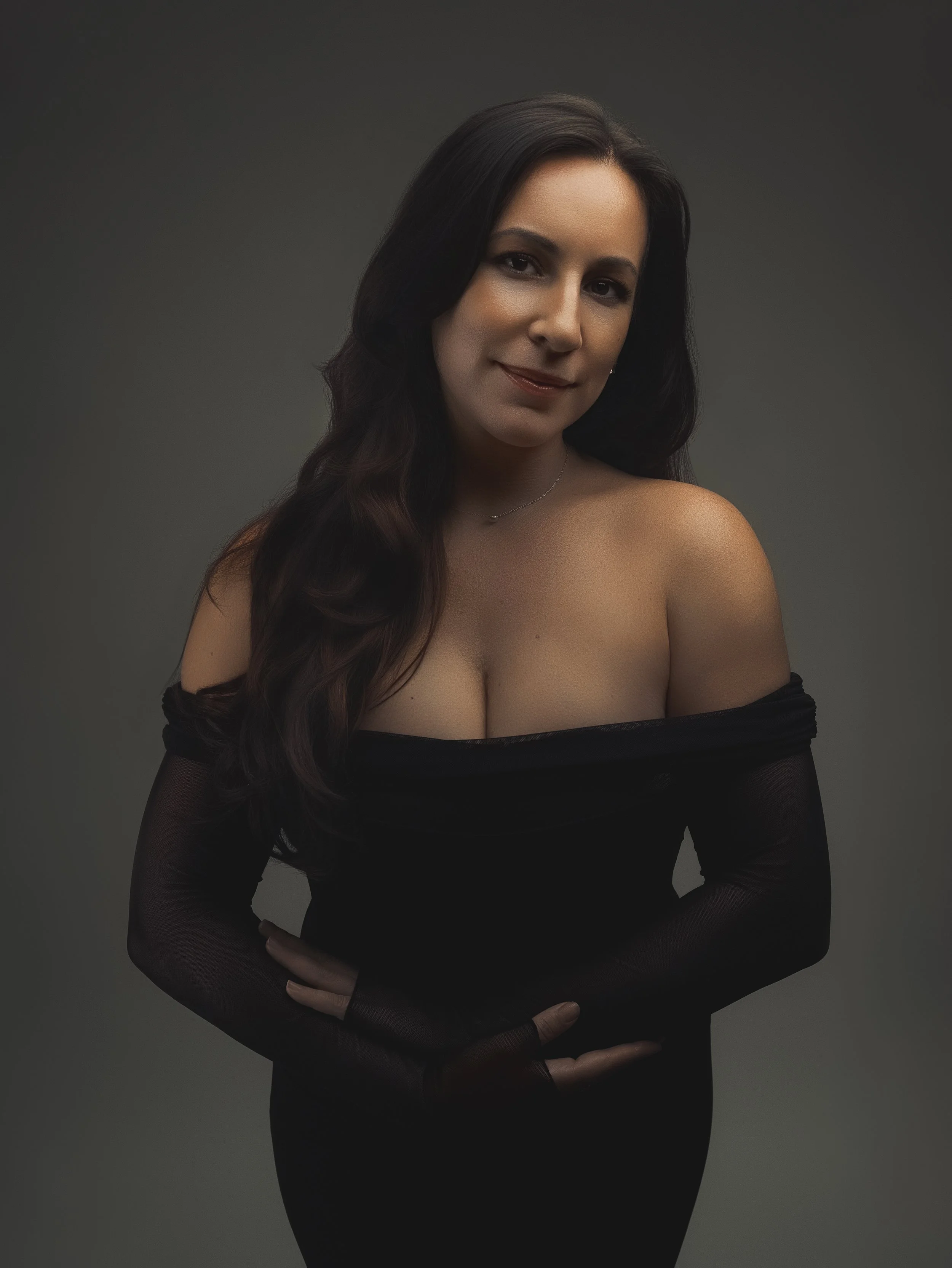 A woman with long dark hair, wearing a black off-shoulder dress and black gloves, posed against a dark background