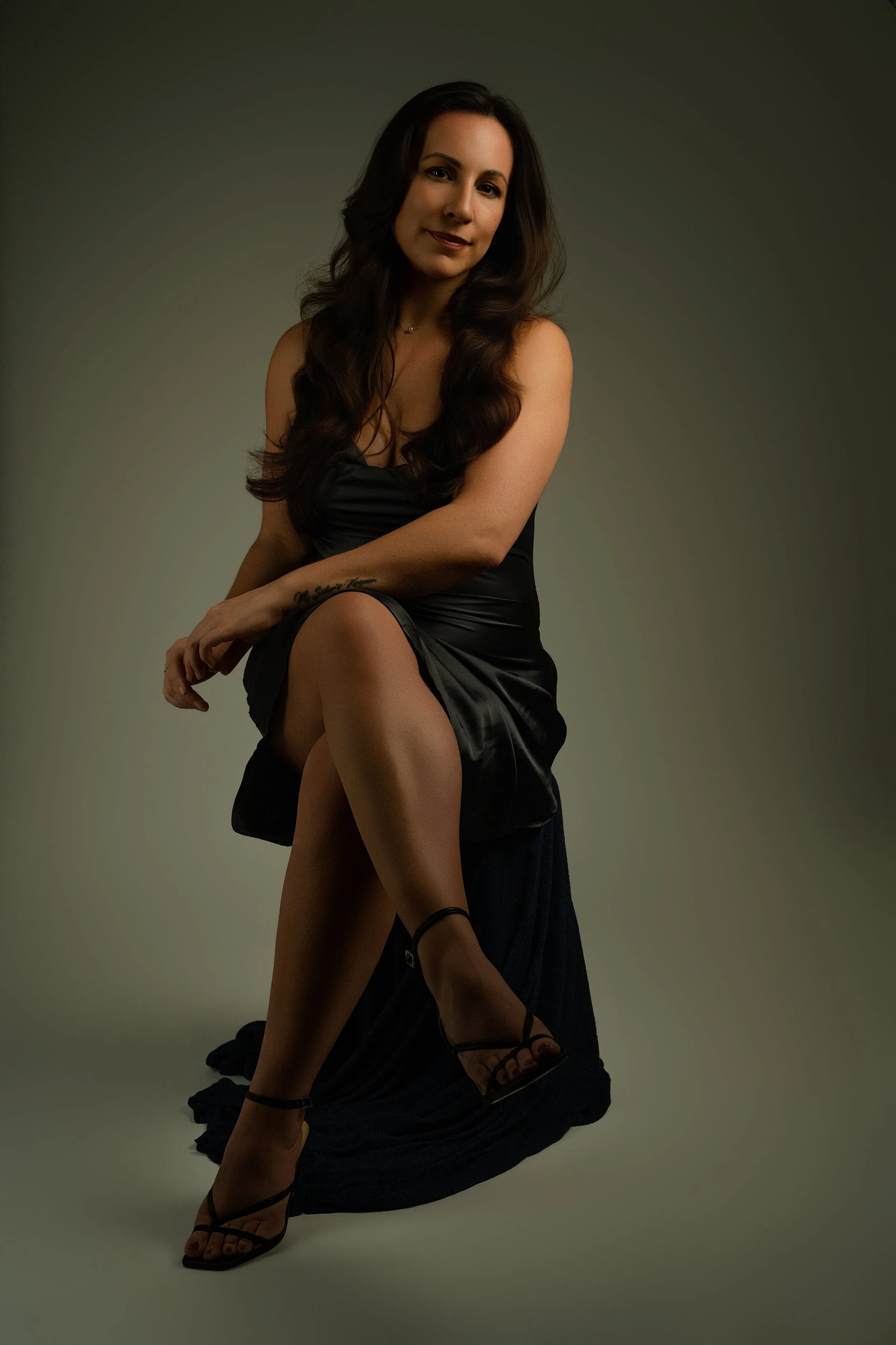 A woman with long, dark, wavy hair wearing a black satin dress and black high-heeled sandals, sitting on the floor with a light background.