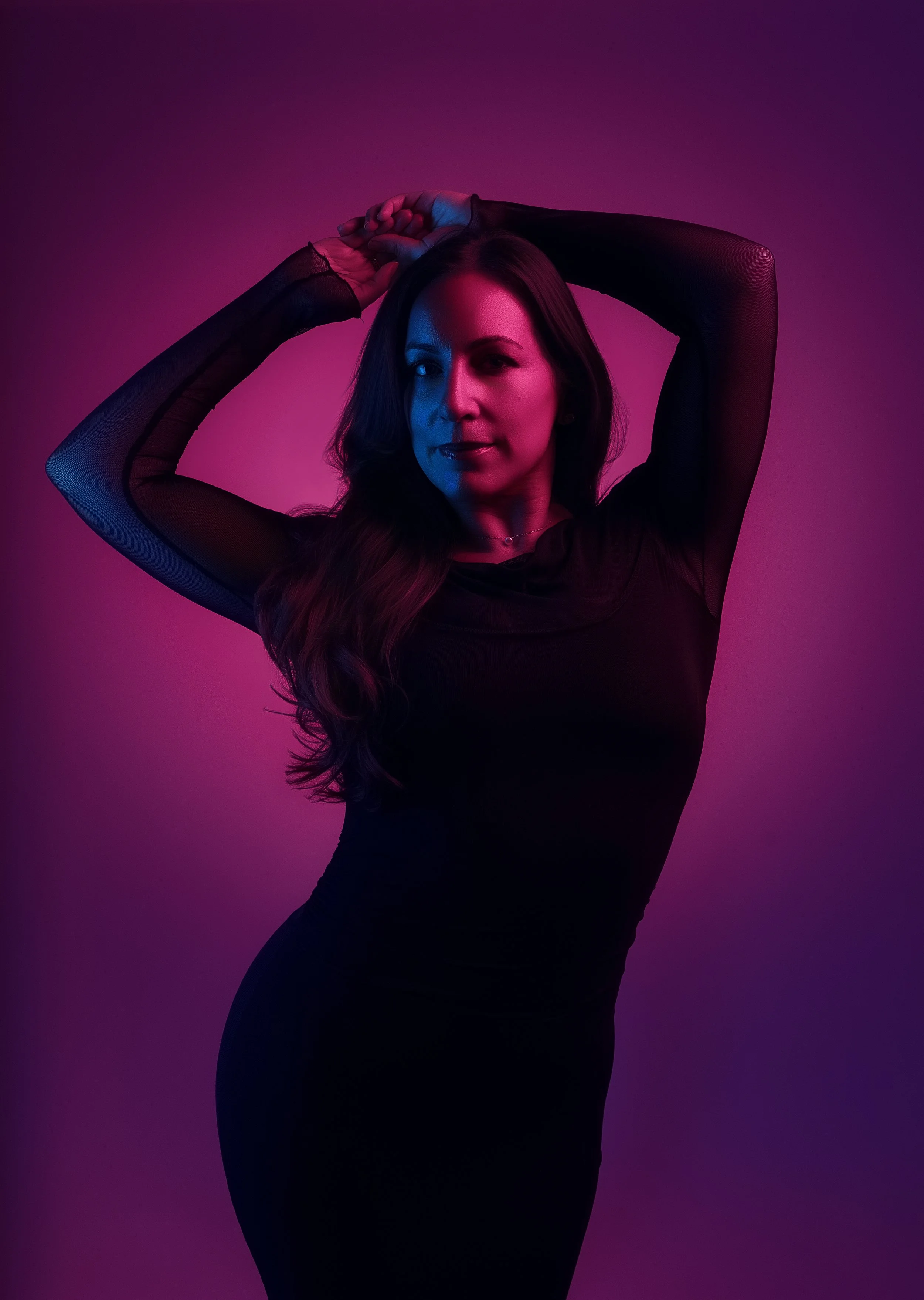 A woman with long hair wearing a black dress and sheer sleeves, posing with her hands above her head against a purple-pink gradient background.