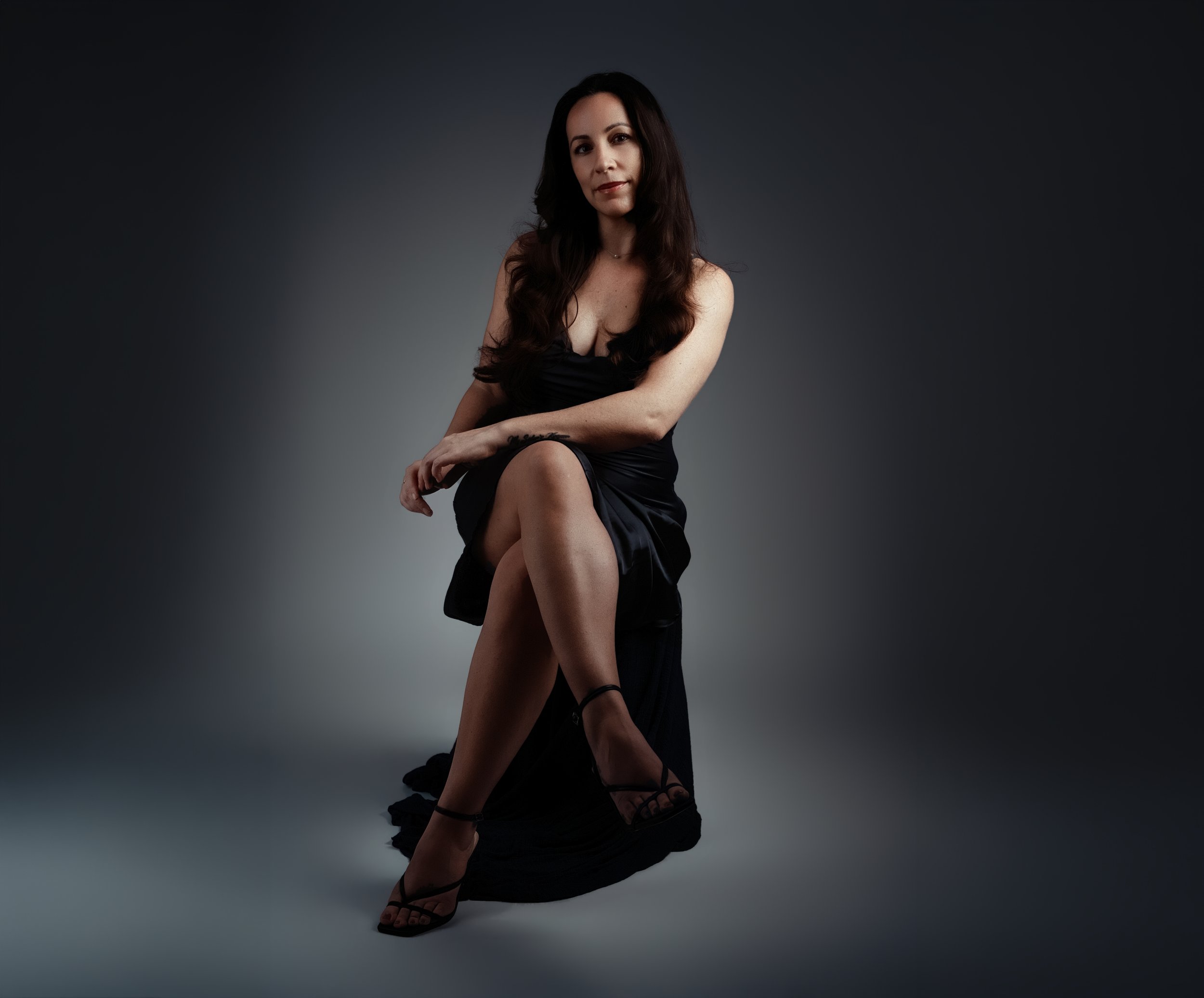 A woman with long dark hair wearing a black dress and heels, sitting on a dark floor against a gray background.