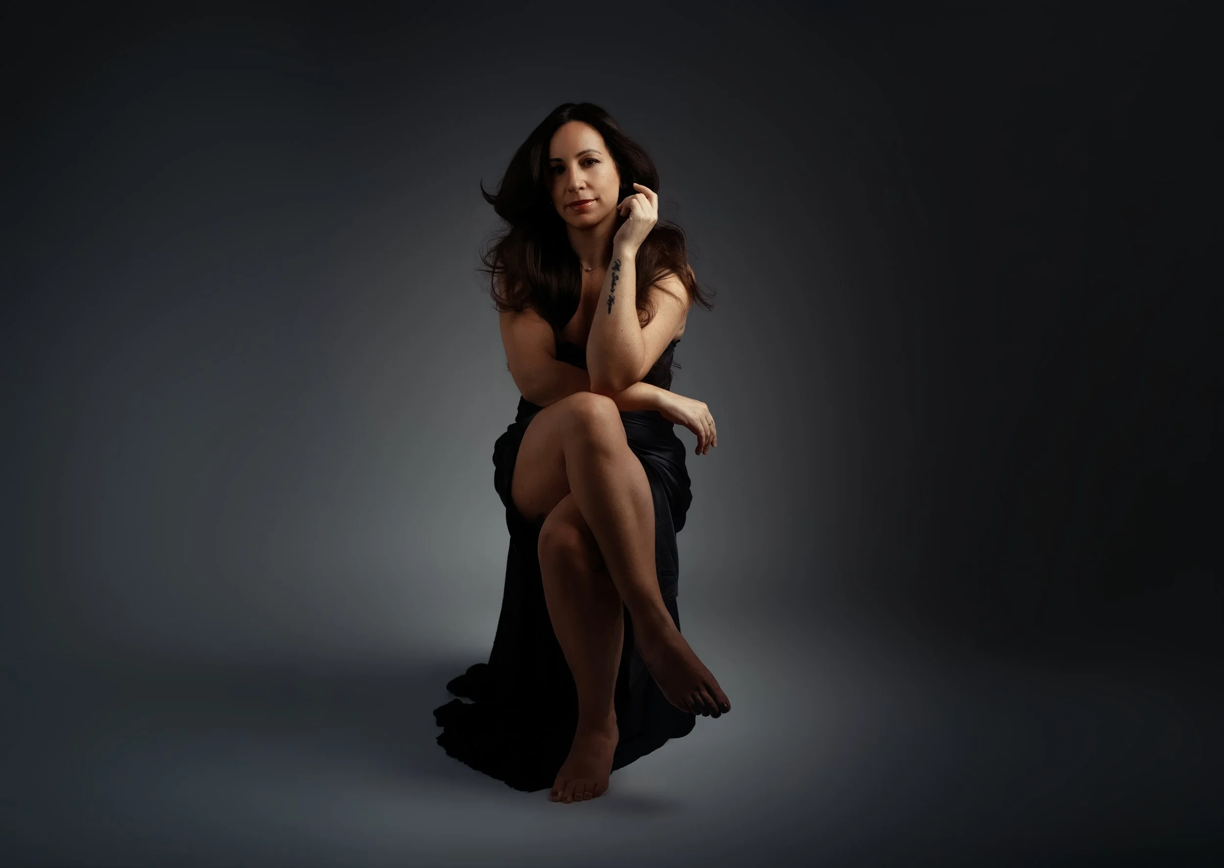 A woman sitting on a black fabric, with one leg crossed over the other, against a gradient gray background. She has long dark hair, a tattoo on her left arm, and is dressed in a black outfit.