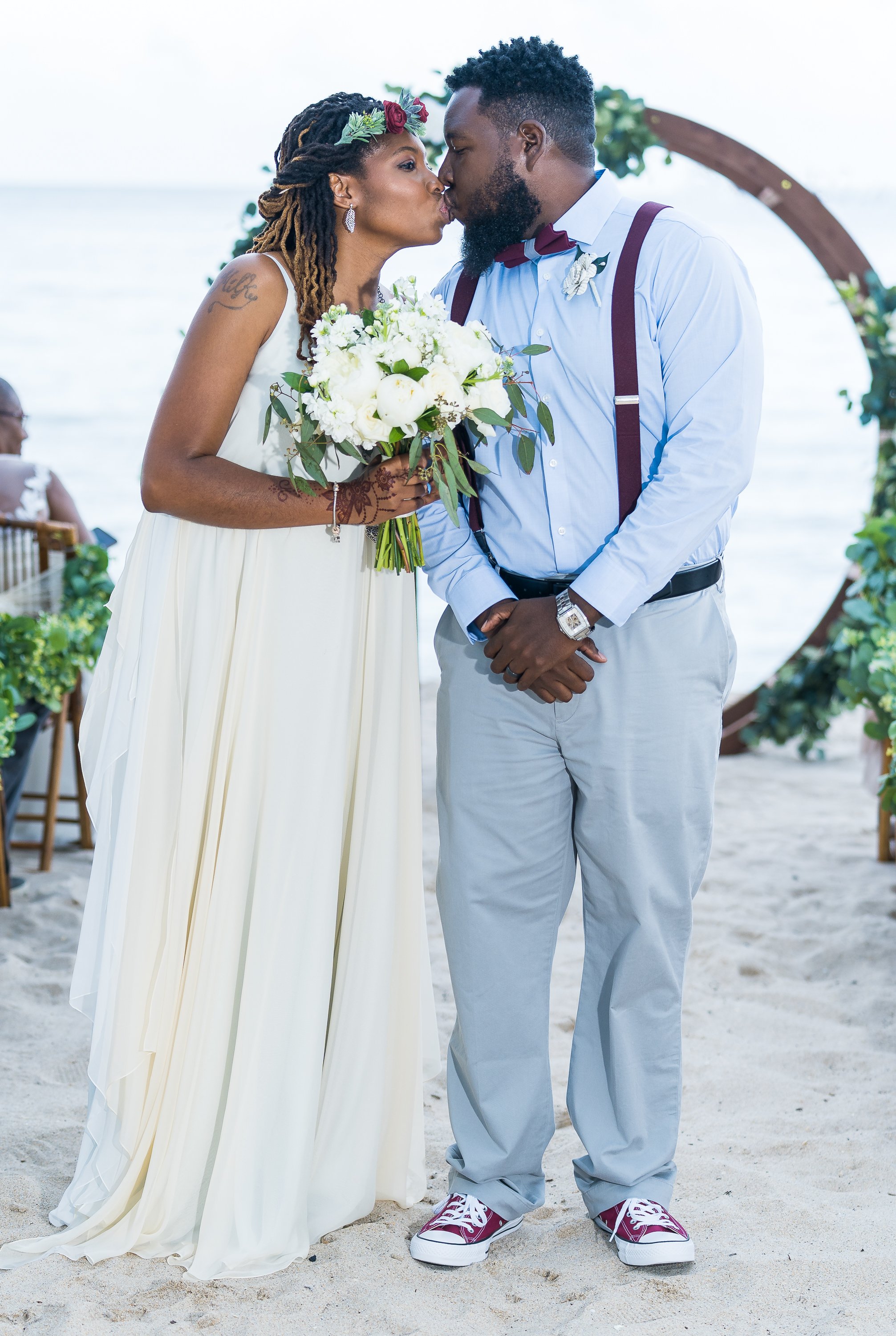 Bride and groom kissing on a sandy beach, with the bride holding a bouquet and the groom wearing a blue shirt and red bow tie.