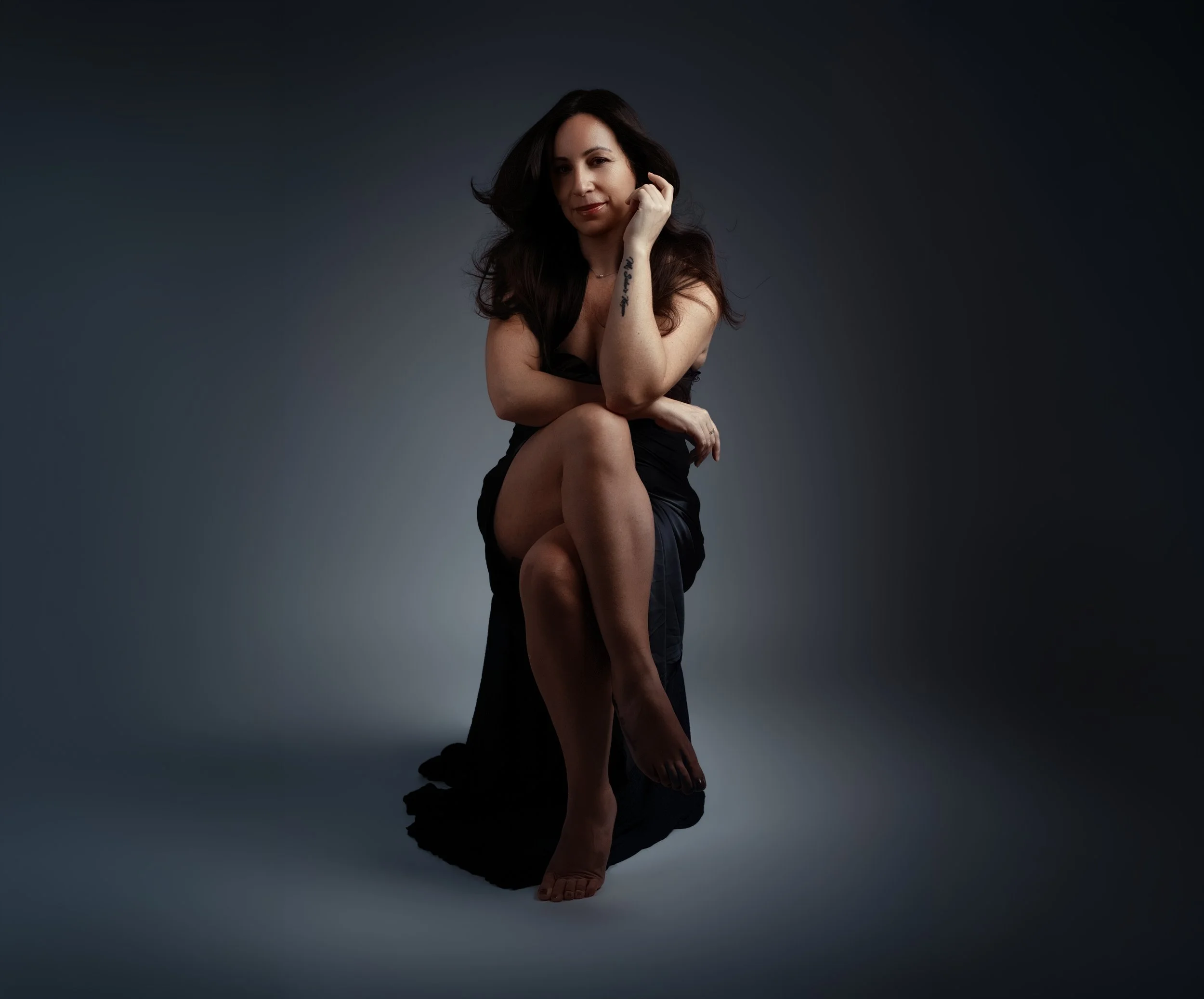 A woman with long dark hair sitting on the floor with her legs crossed, wearing a black dress, in a studio with a dark gradient background, looking directly at the camera.