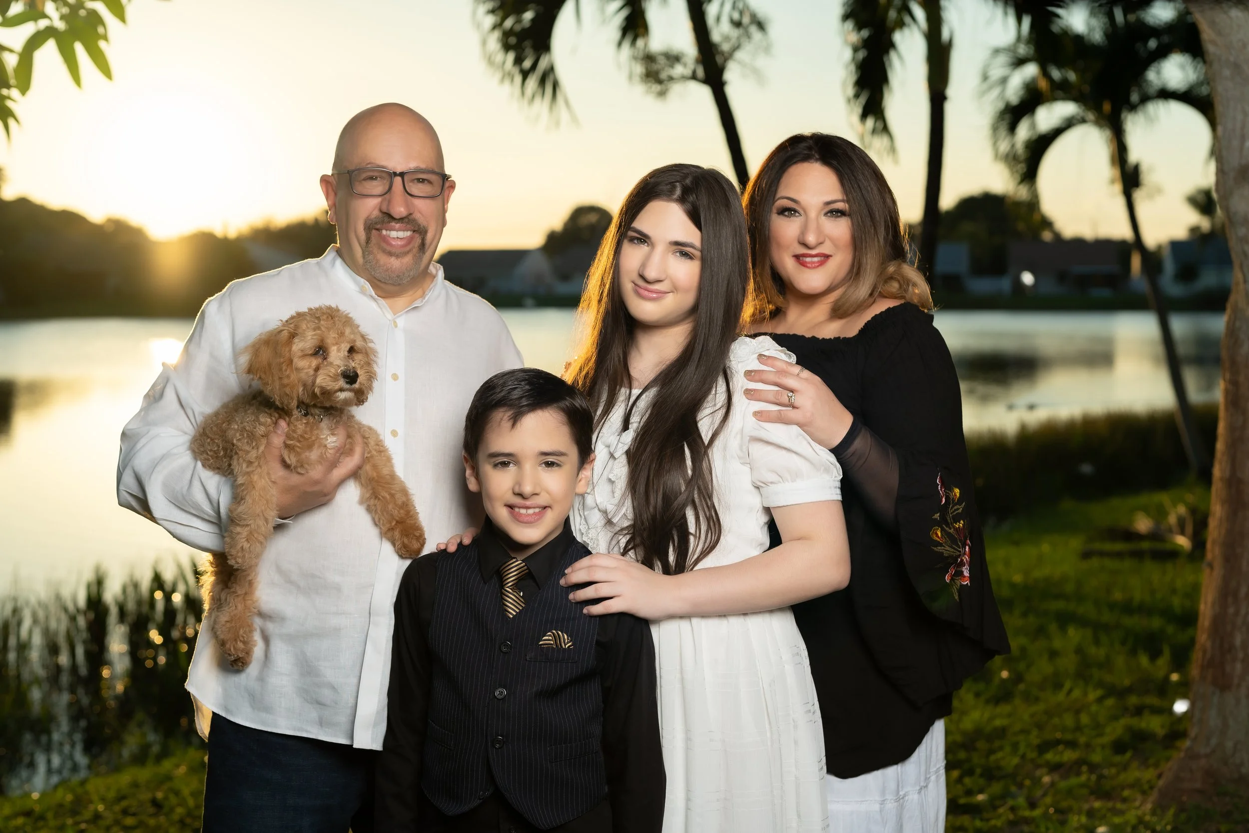 Family portrait with four people and a dog outdoors near a lake at sunset, featuring palm trees in the background.