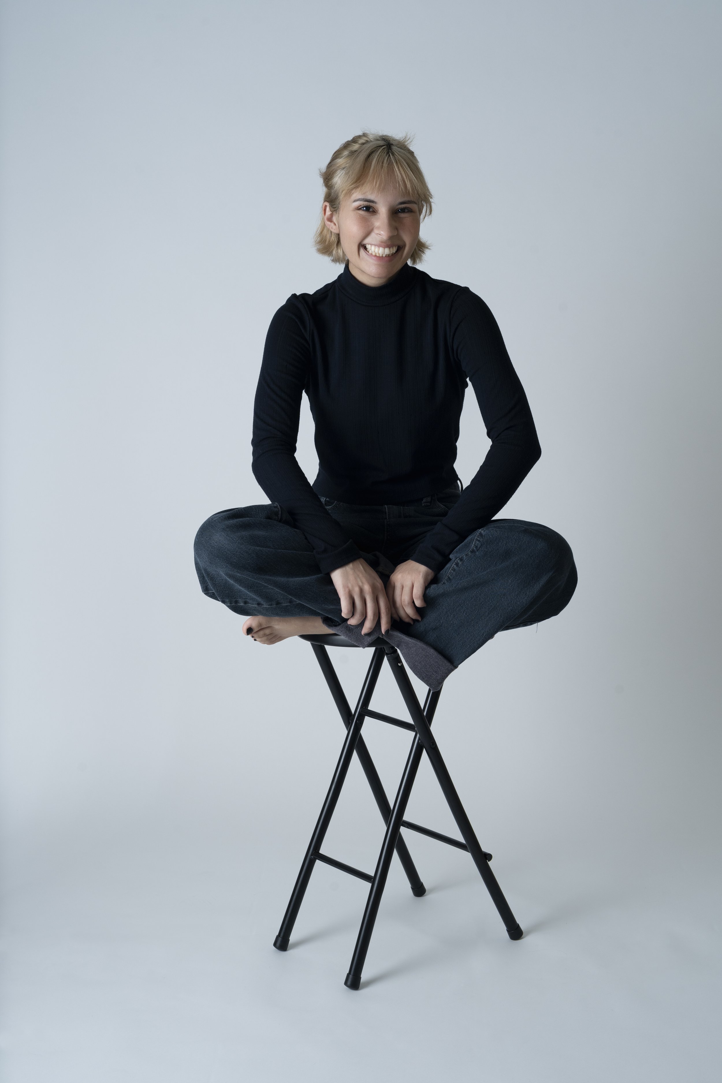Person sitting cross-legged on a stool, wearing a black turtleneck and jeans, smiling, with short blond hair.