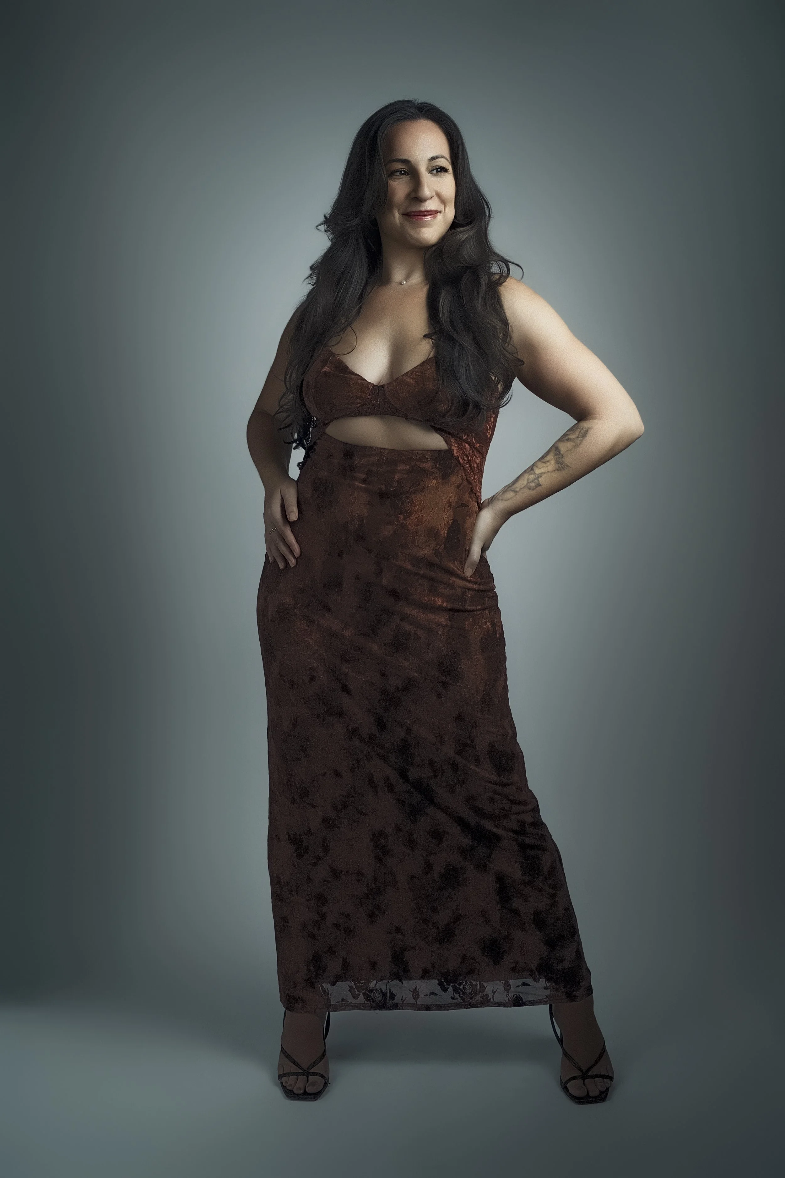 A woman with long dark hair wearing a brown velvet evening gown with a cutout, standing with one hand on her hip and smiling.