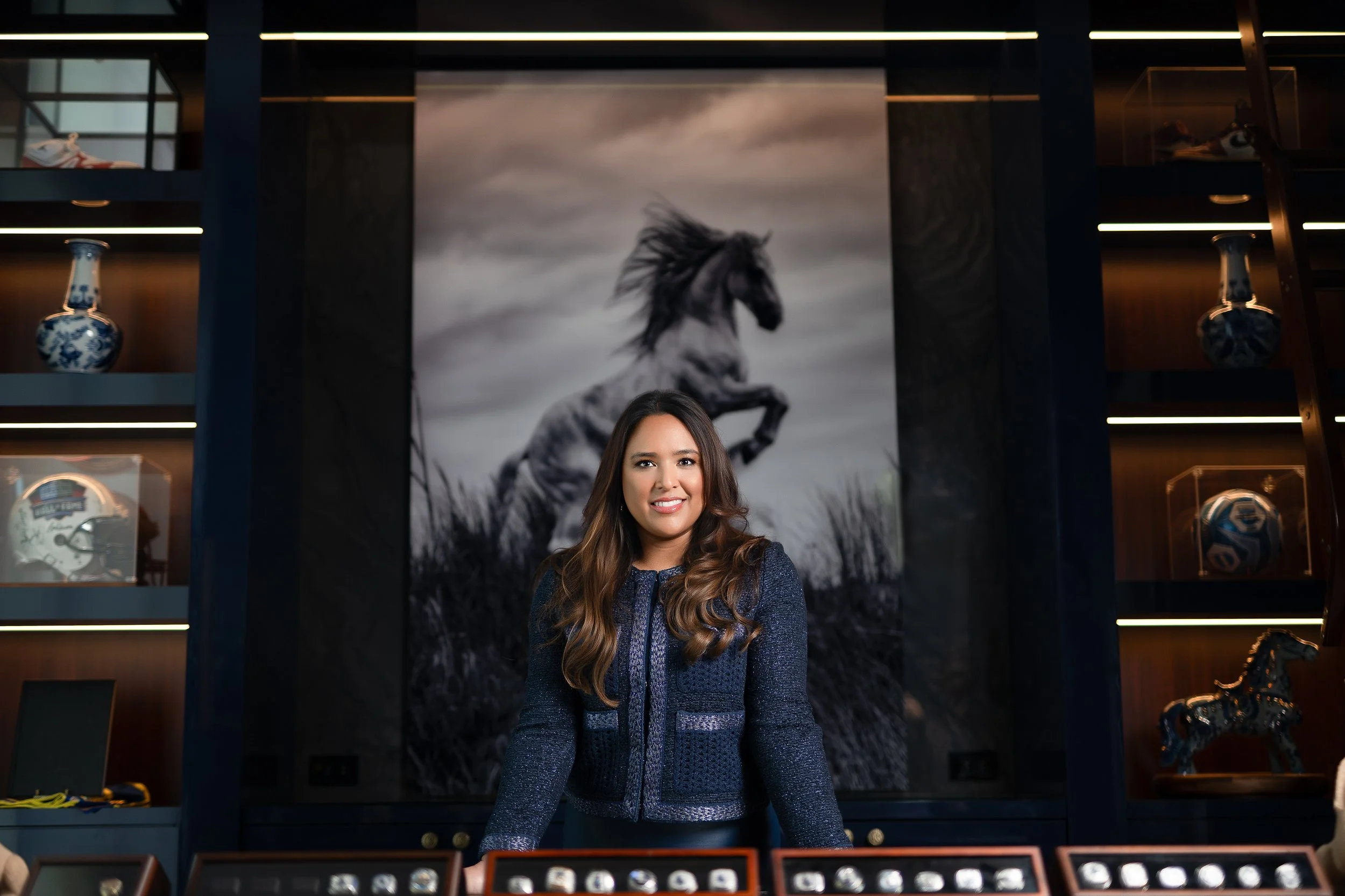 A woman with long wavy brown hair smiling in front of a large black-and-white mural of a rearing horse, surrounded by display shelves with decorative vases, a football, and a horse figurine.